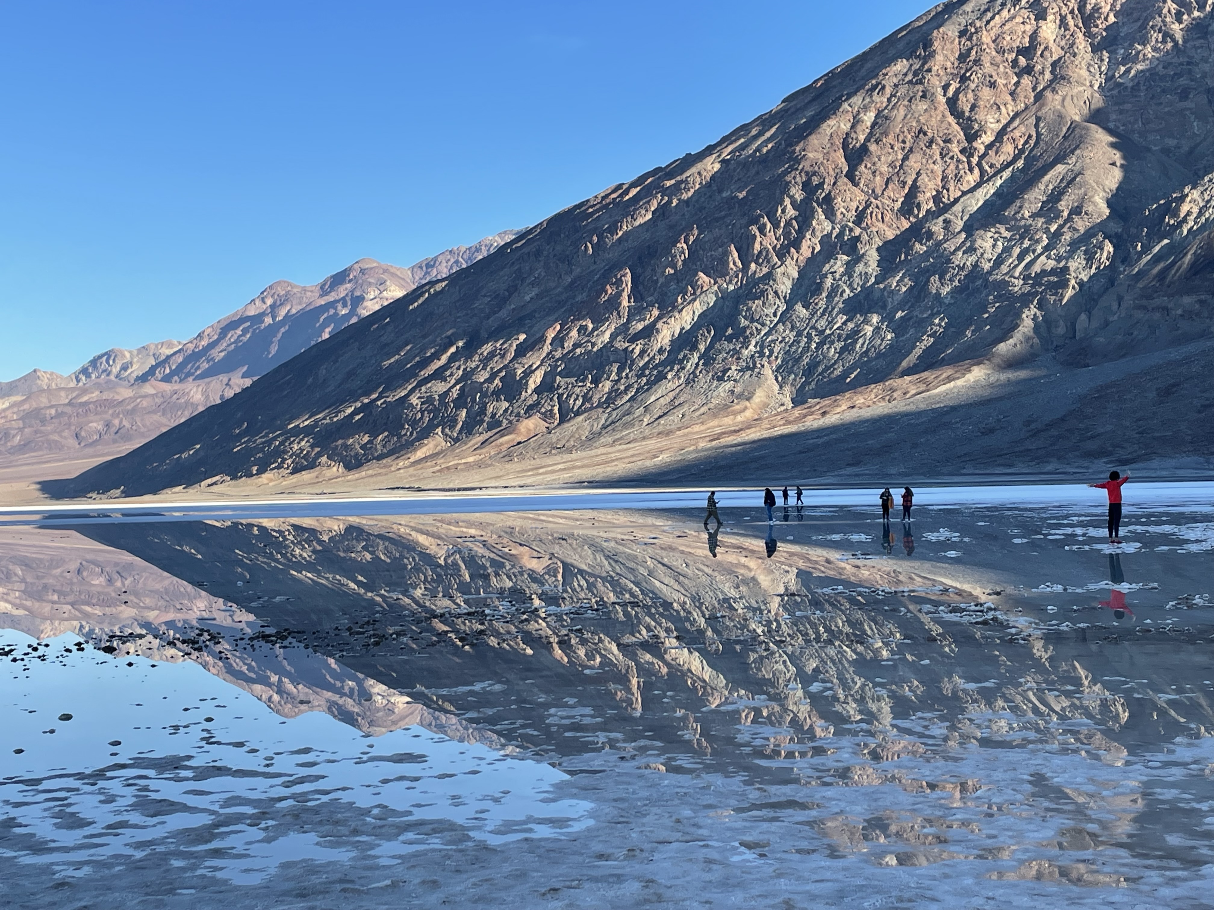 A brown hillside slopes up to the right. It is reflected upside-down in calm water. Several people stand in the distance in the water.