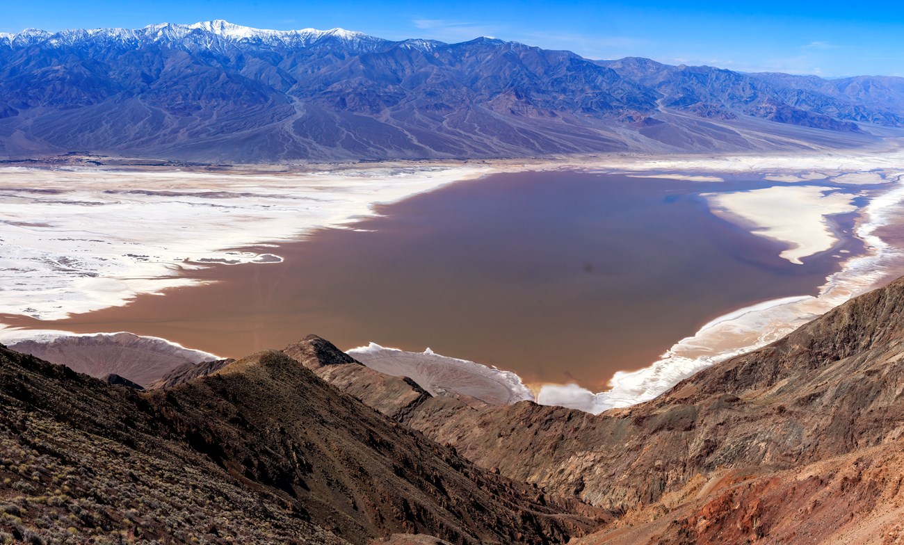 View from a mountain looking down at a wide, flat valley with a brown lake in it.