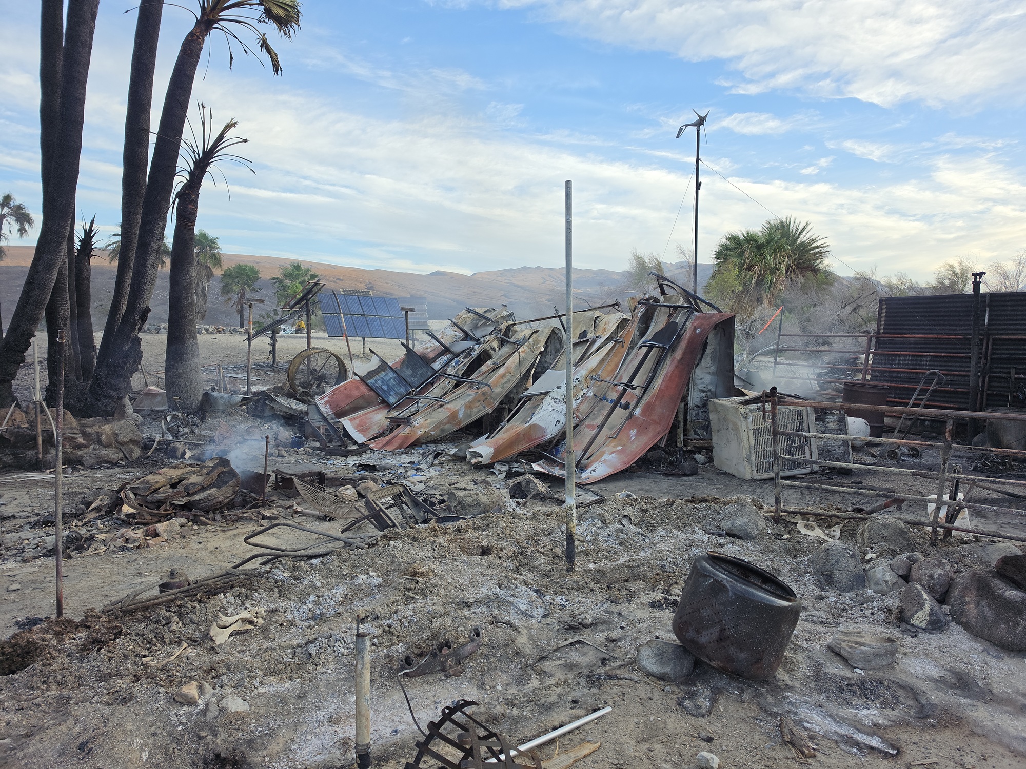 Dark-colored burned palm trees on the left. Orange and gray metal debris in in the center and foreground, along with two narrow vertical poles.