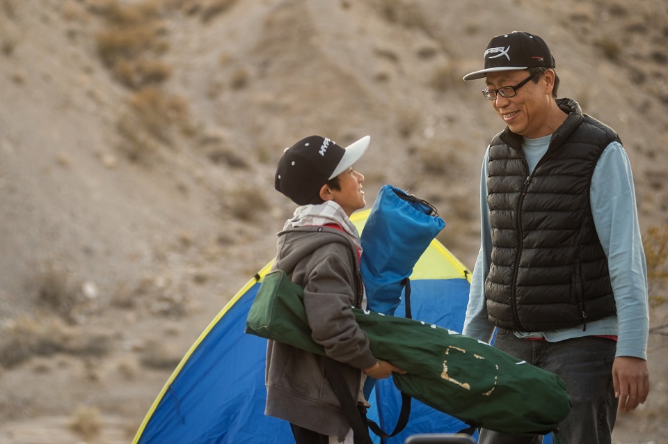A boy carrying two camp chair bags looks up at a man. Their tent is in the background.