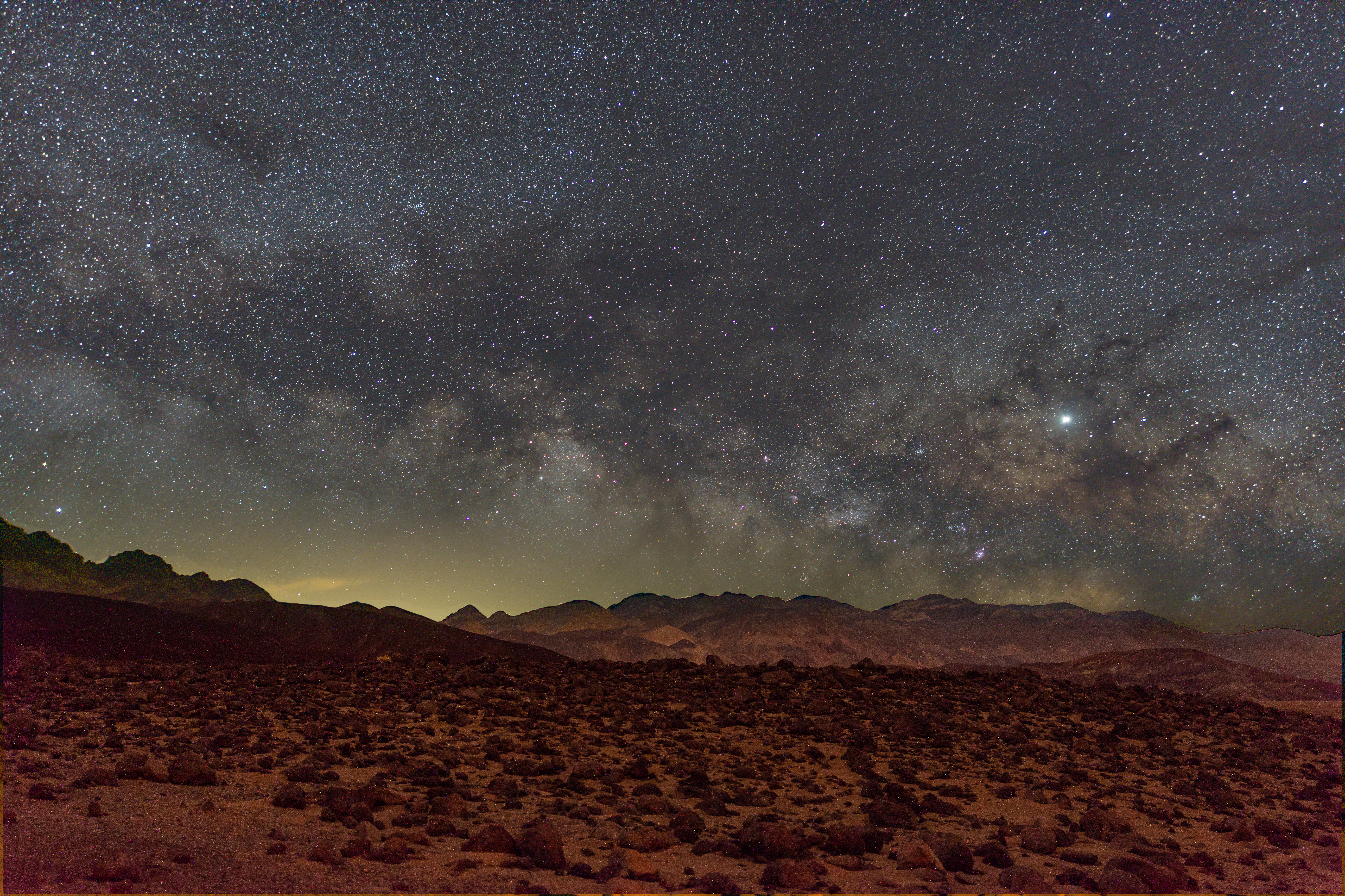 Hundreds of stars dot the sky, forming what looks like a white haze of distant stars  horizontally across the skyline. The foreground is slightly reddish with small shrubs.