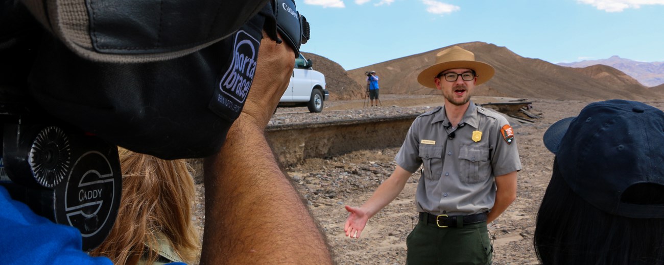 Ranger in uniform speaking to a camera in from of brown mountain landscape.