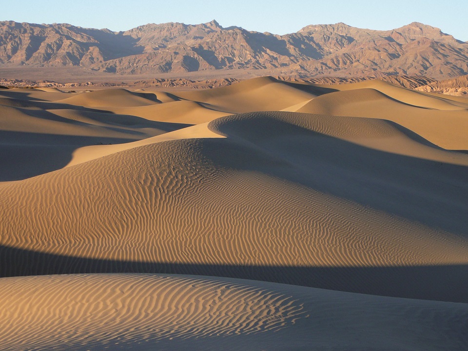 Mesquite Flat Sand Dunes Tan-colored sand dunes are in the foreground, with rocky mountains and a cloudless sky in the background.