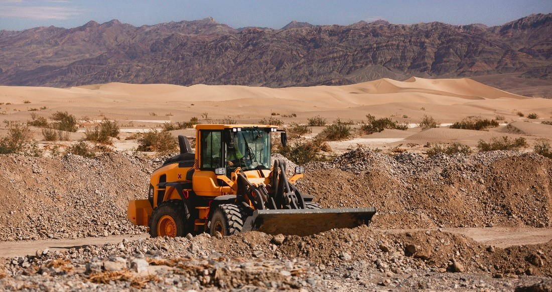 A yellow tractor moves brown dirt and rocks from a parking lot with sand dunes in the background. Behind the sand dunes is a mountain landscape.