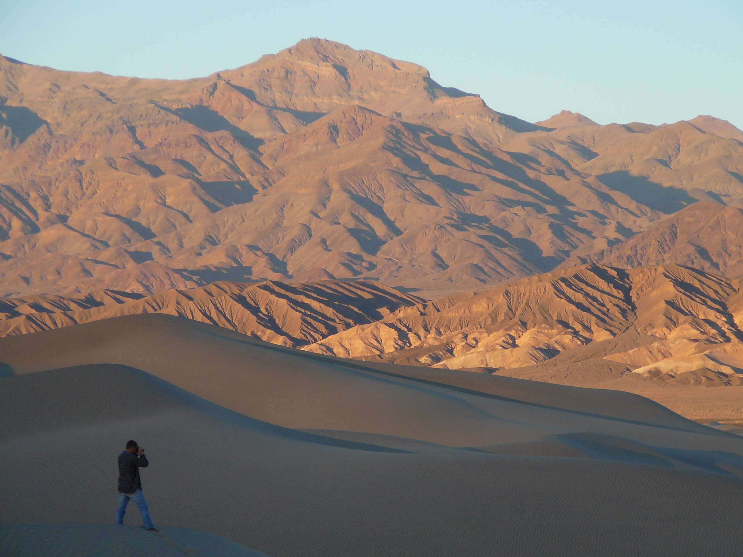 A desert mountain range in the background looks red-brown in late afternoon light. A single person stands with a camera on a sand dune in the foreground.