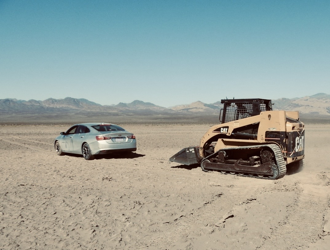 A yellow skid steer and a light gray car are on a flat dirt surface with vehicle tracks. Mountains are visible in the distance.