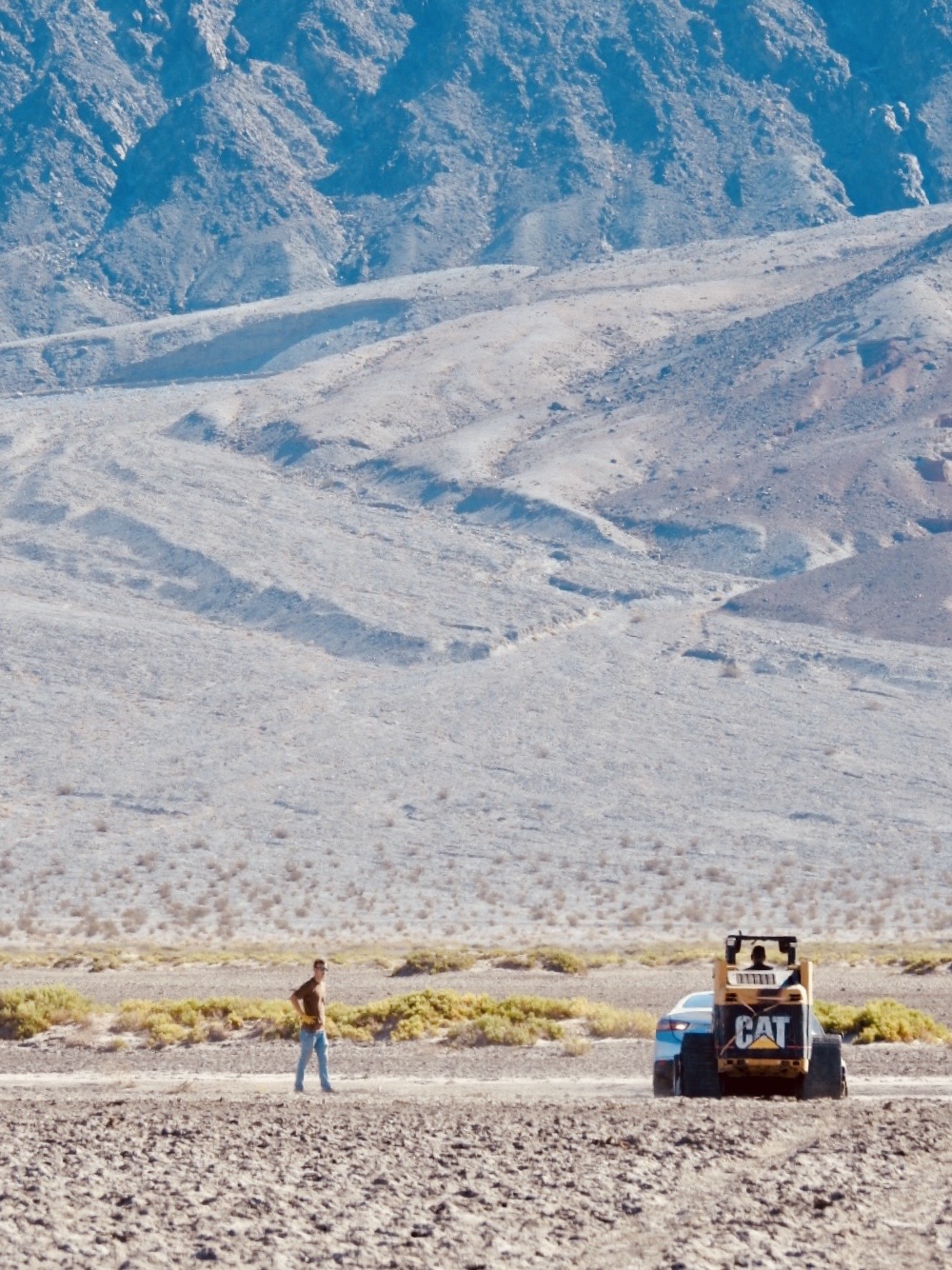 A person (on left) and a skid steer ("CAT" written on back) and car are on flat brown ground. Brown mountains are in background.