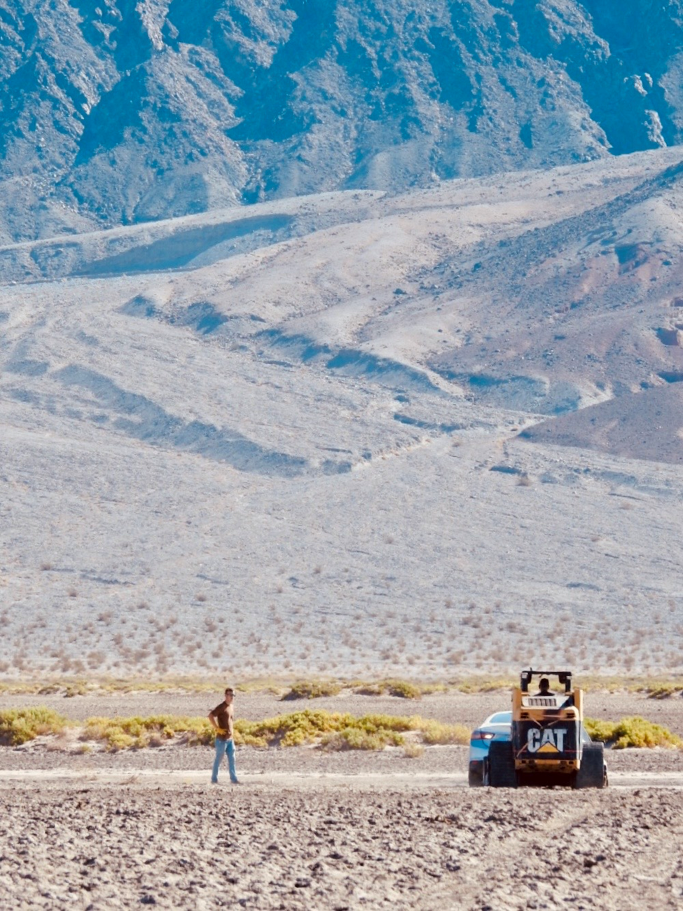 A person (on left) and a skid steer ("CAT" written on back) and car are on flat brown ground. Brown mountains are in background.