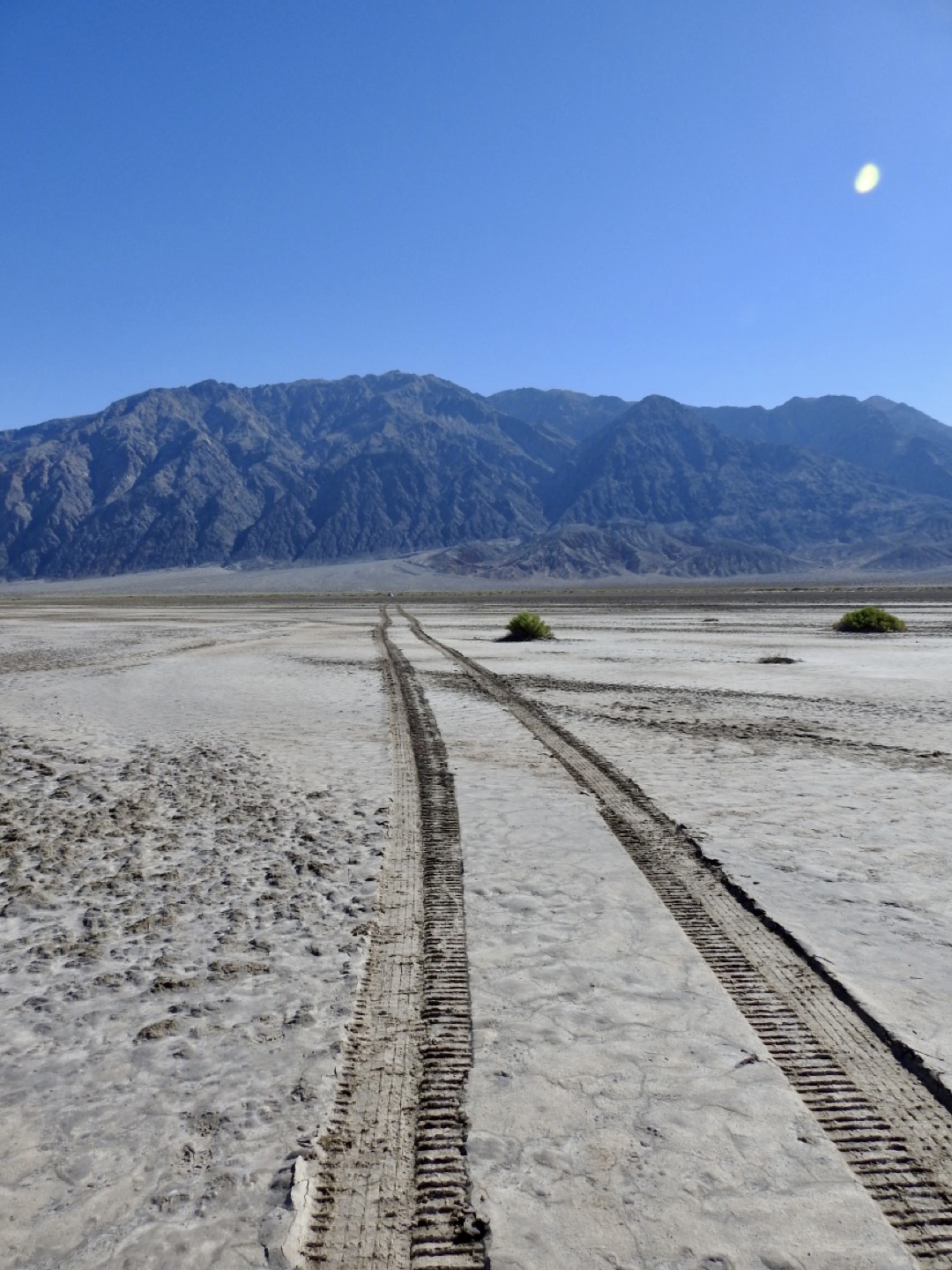 Vehicle tracks run from the camera out into a flat open ground toward distant mountains.