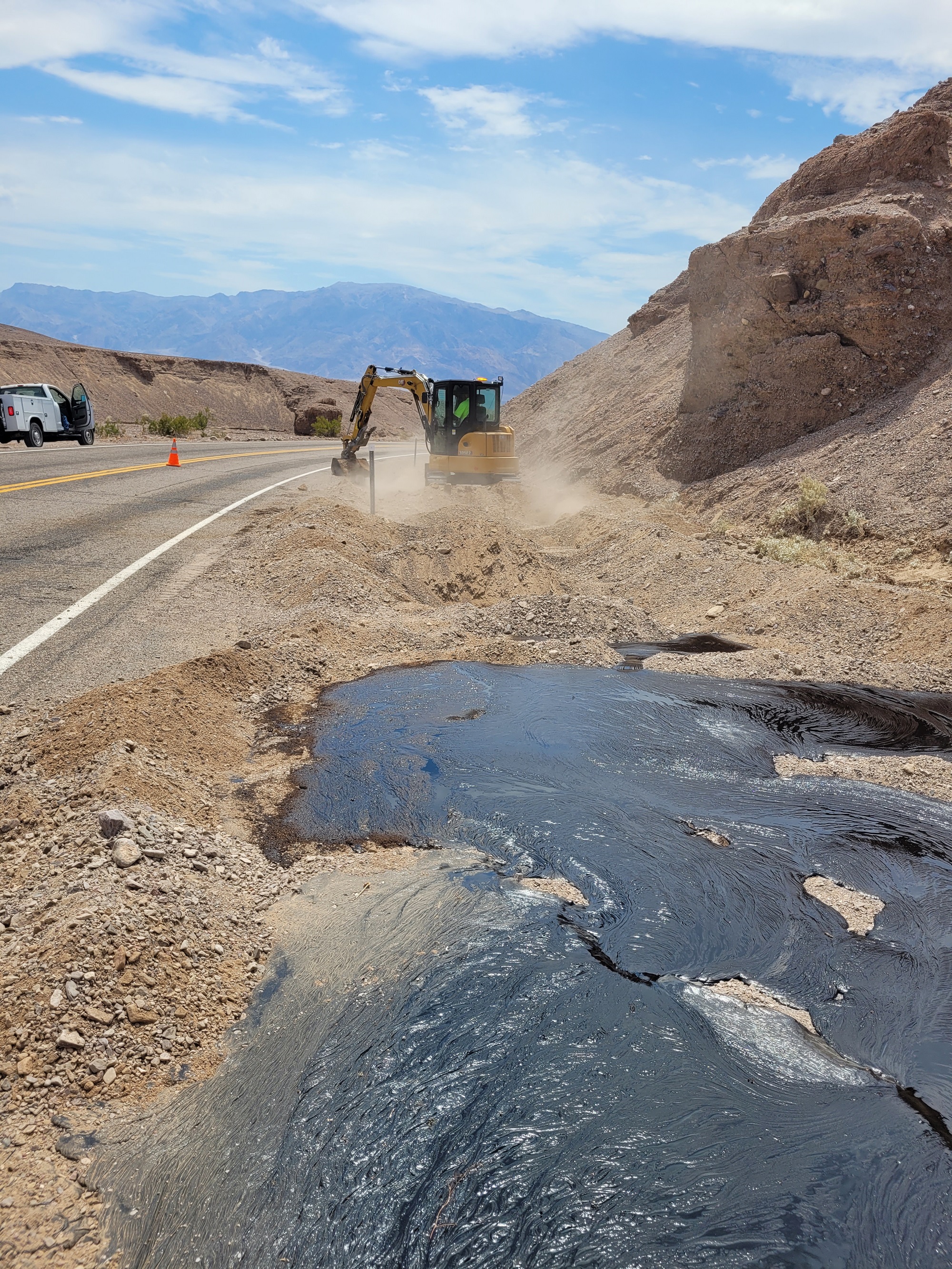 Black thick liquid in the foreground and a yellow machine in the background next to a paved road.