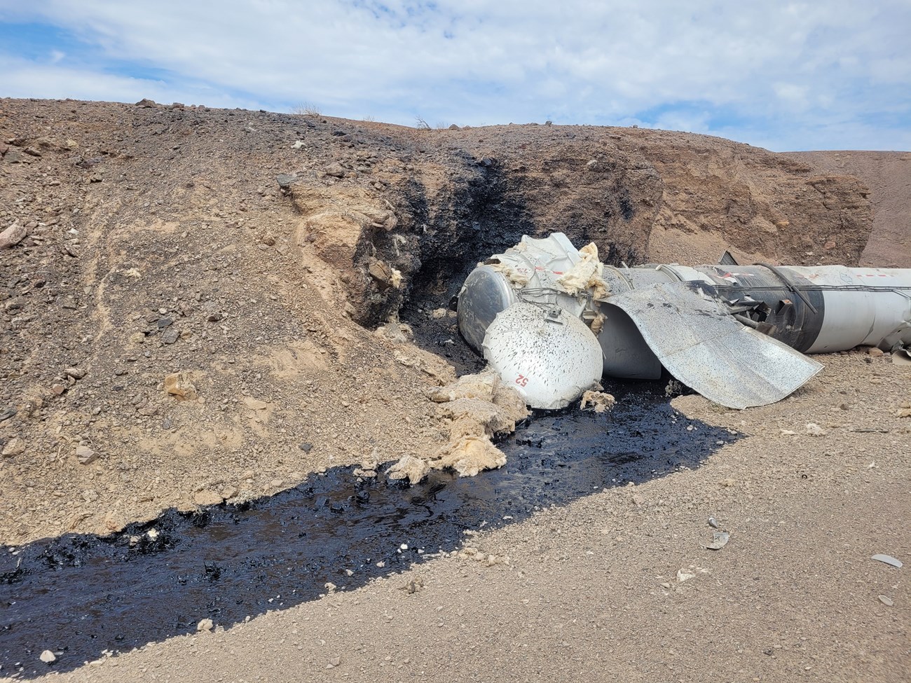 A wrecked trailer tank on the right side with a wet, black stream out of it on the ground.