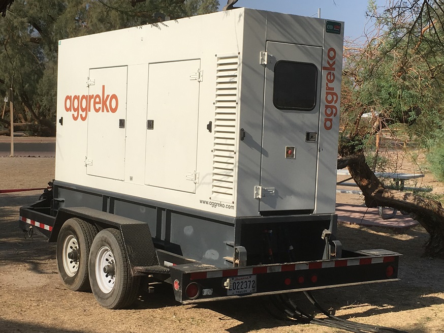 A white trailer-mounted generator is hooked by thick cables to an electrical panel behind Furnace Creek Visitor Center.