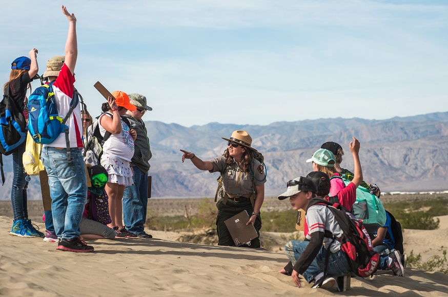 A park ranger kneels in sand while 4th grade students raise their hands to answer a question