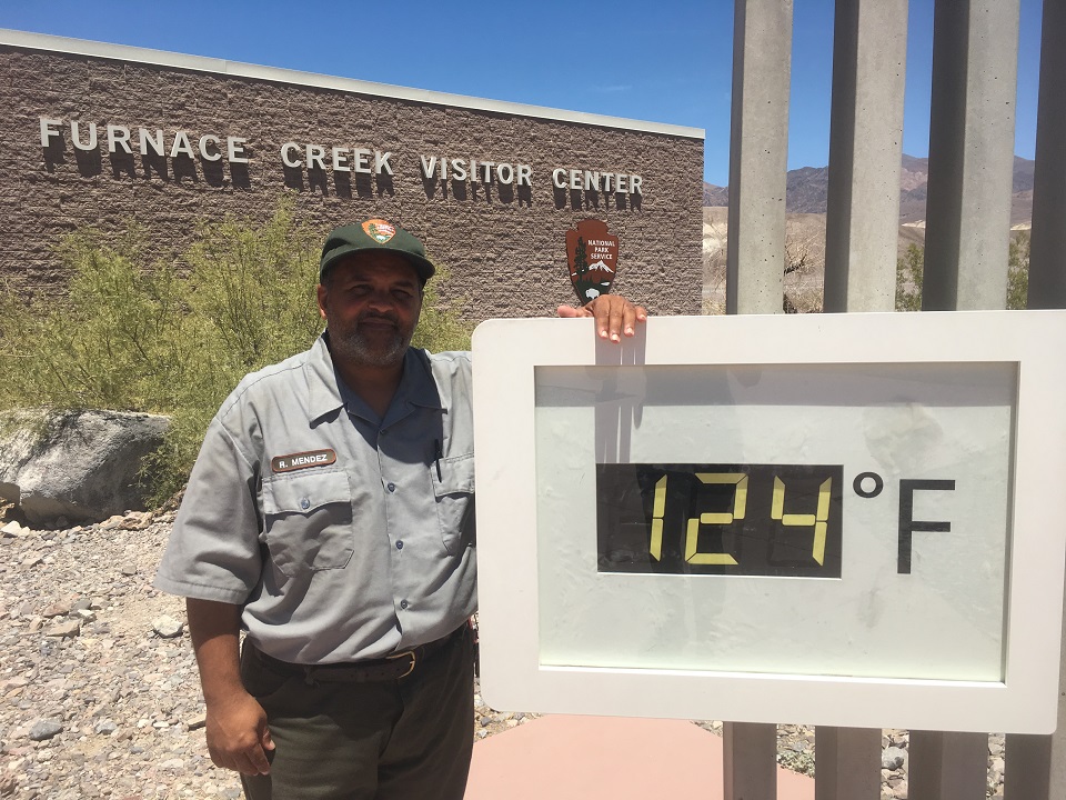 Park ranger Roberto Mendez stands next to the temperature reading 124 degrees Farenheit. Furnace Creek Visitor Center and the National Park Service arrowhead can be seen in the background.