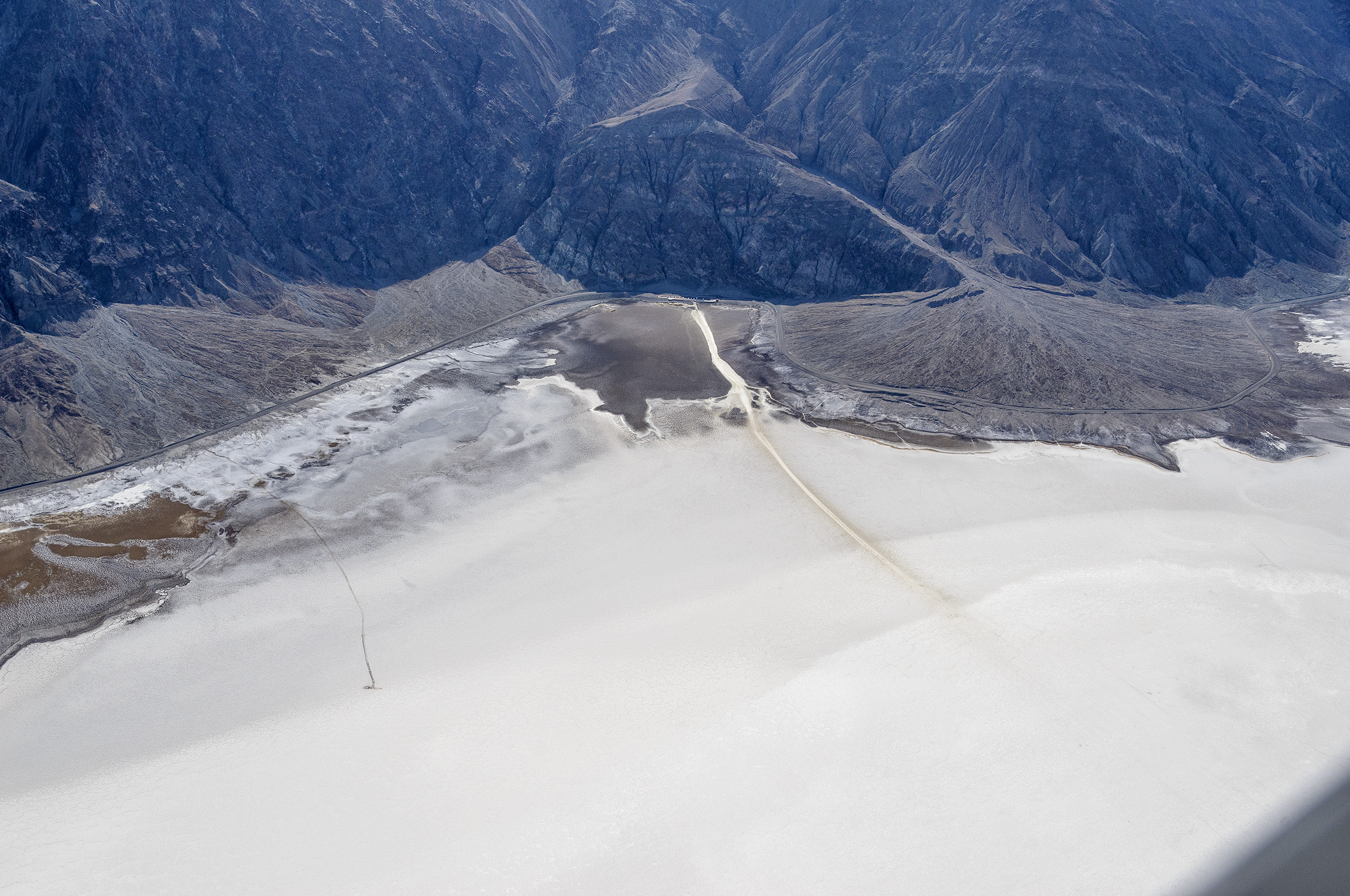 Photograph taken looking down from an airplane shows a broad white salt flat, a trail, road, and the side of dark mountains.