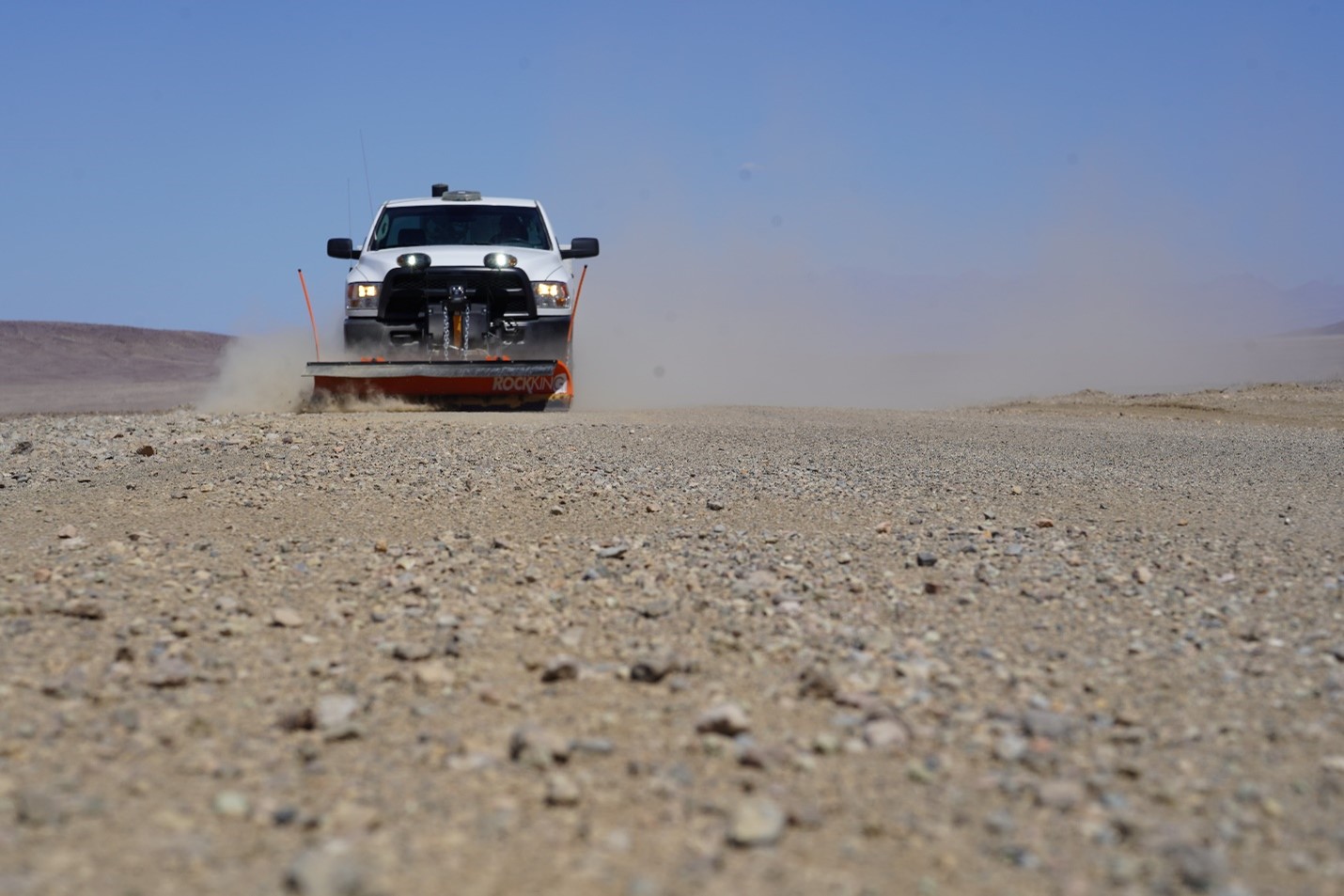 Photo taken from ground level showing gravel on a paved road and a white truck with a plow clearing gravel.