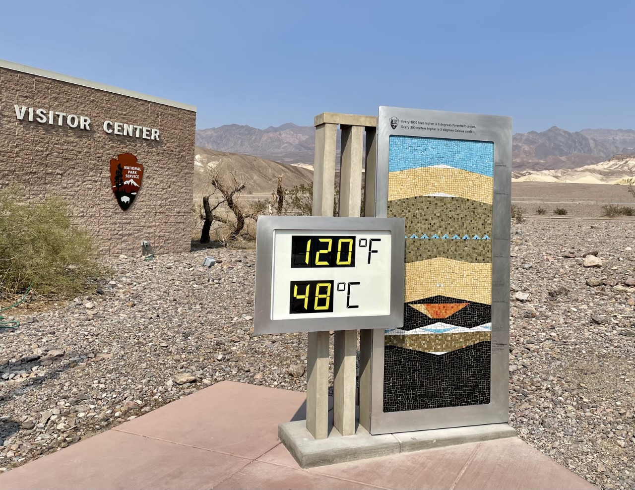 Outdoor digital thermometer display of 120F, 48C. A colorful ceramic mosaic approximately 6 ft tall depicting a valley, crater, charcoal kilns and mountains is to the right of the thermometer. Background: bulding with "Visitor Center" and NPS Arrowhead