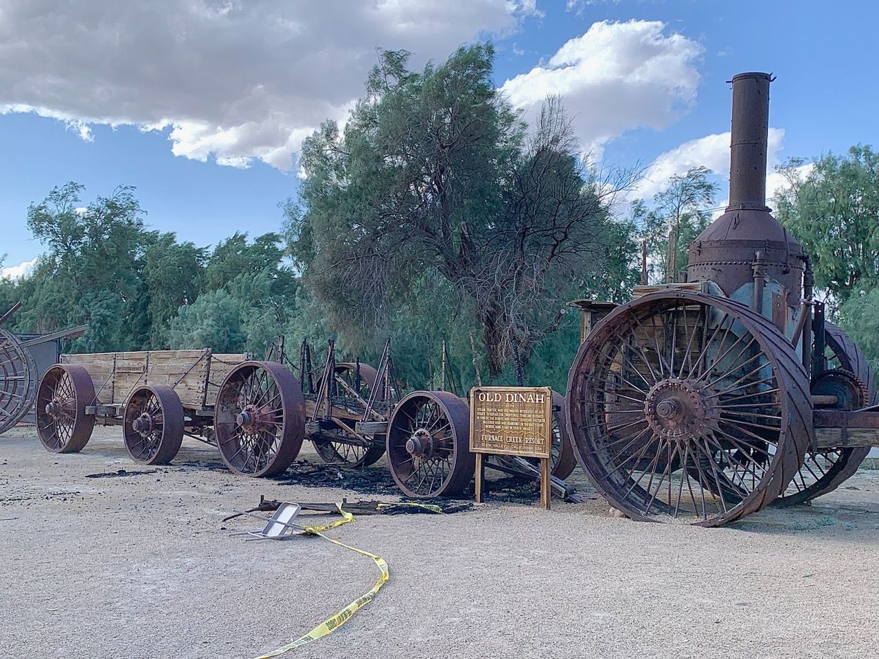 A historic borax wagon and "Old Dinah," a historic steam engine with a burned historic borax wagon in between them and green tamarisk trees in the background.
