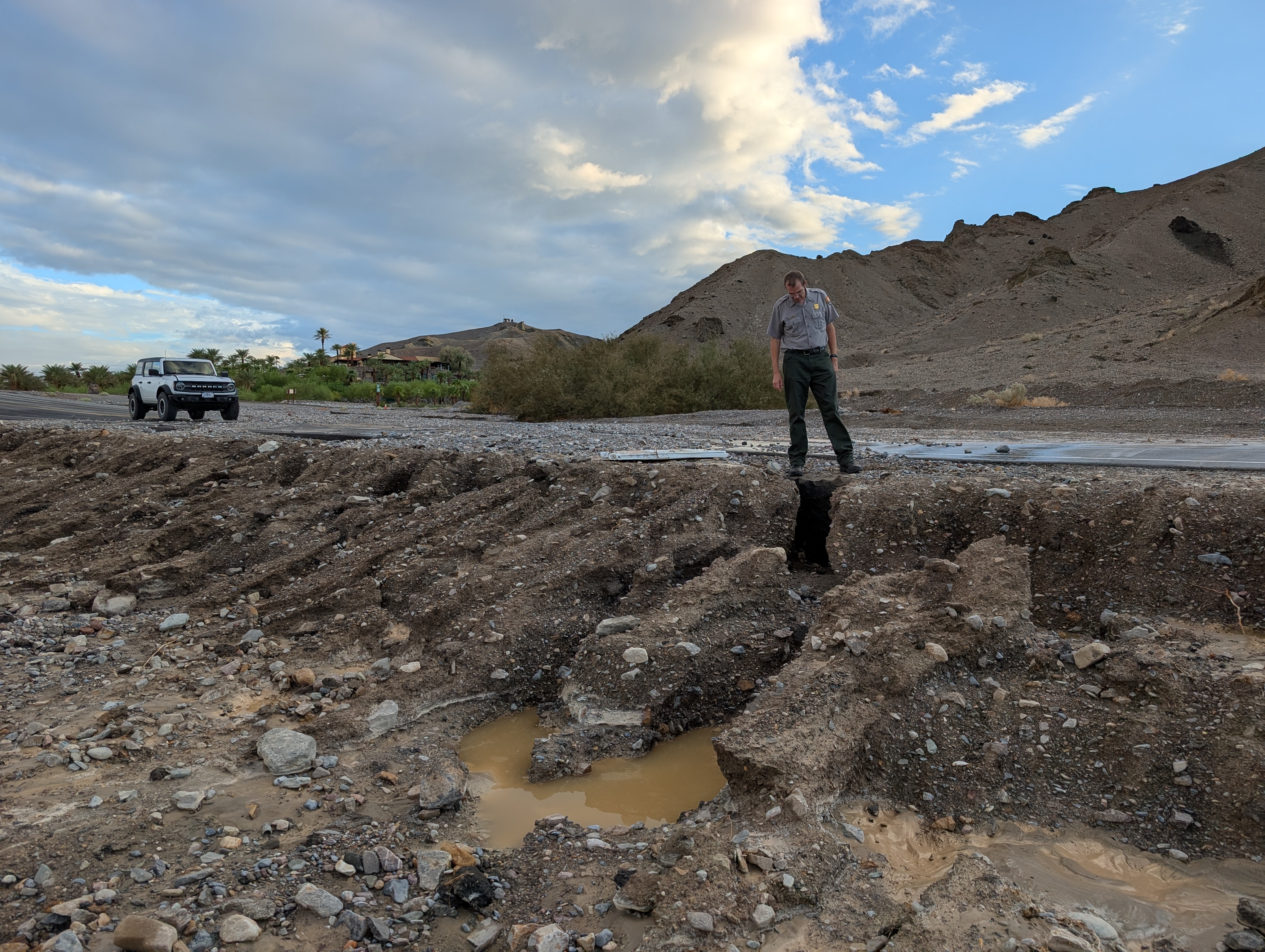 A man in uniform stands on a paved road looking at an eroded shoulder below him.
