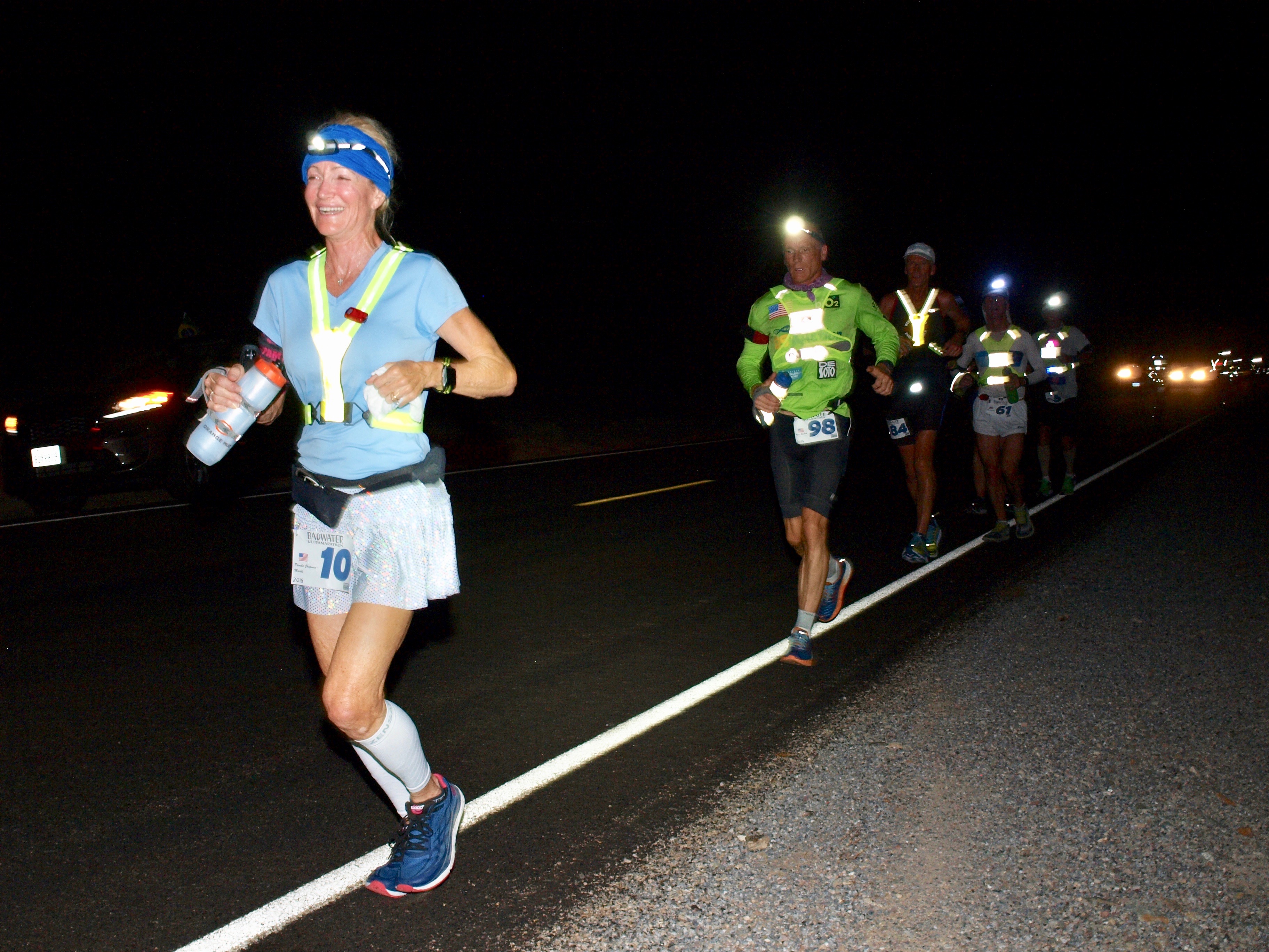 Runners, at night, in a string, run along a paved road.