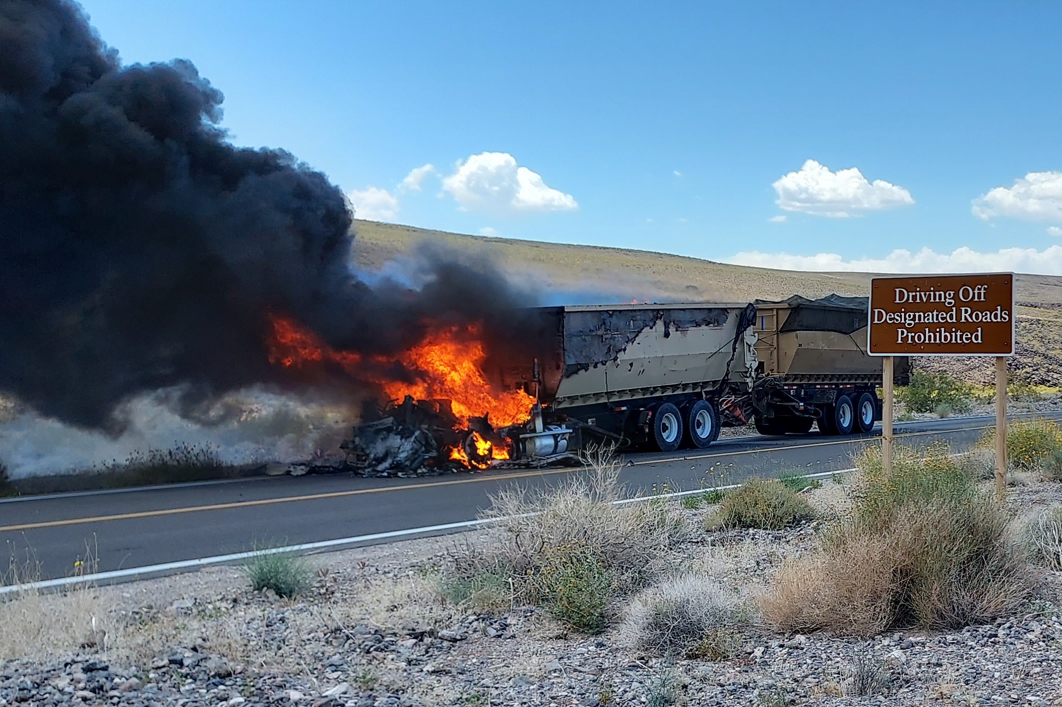 Black smoke billows out from bright red flames that are fully engulfing the engine and cab of a semi-truck. Two tan trailers are behind it. In the foreground on right is a brown sign, "Driving Off Designated Roads Prohibited".