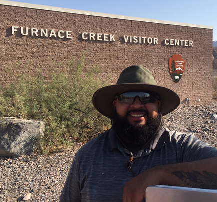 A man wearing sunglasses an a hat stands in front of Furnace Creek Visitor Center.