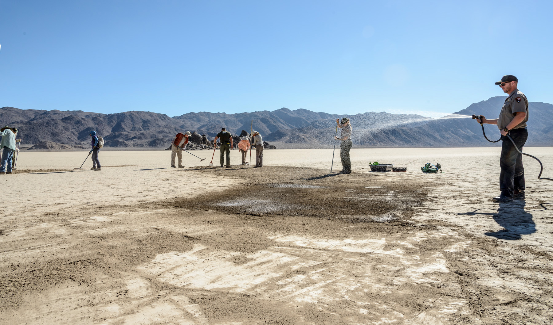 Racetrack Rehab A group of people are raking and watering a large flat playa, where road tracks had been left in the dirt.