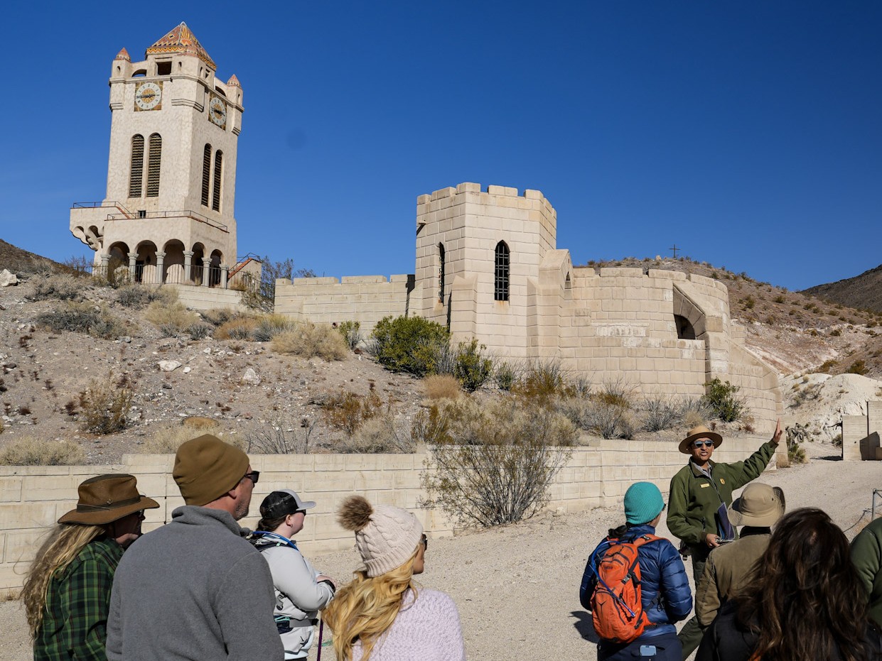 A group of people stand with their backs to the camera facing a man in National Park Service uniform (green jacket and tan flat-rimmed hat). Behind him are two tan-colored buildings, sparsely vegetated hills, and a deep blue sky.