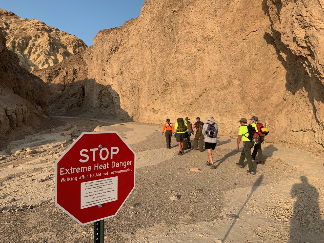 SAR Golden Canyon NPSKRoss IMG_0993 a stop sign saying Extreme Heat Danger hiking after 10 AM not recommended, with brightly colored searchers walking by a wheeled carryout litter