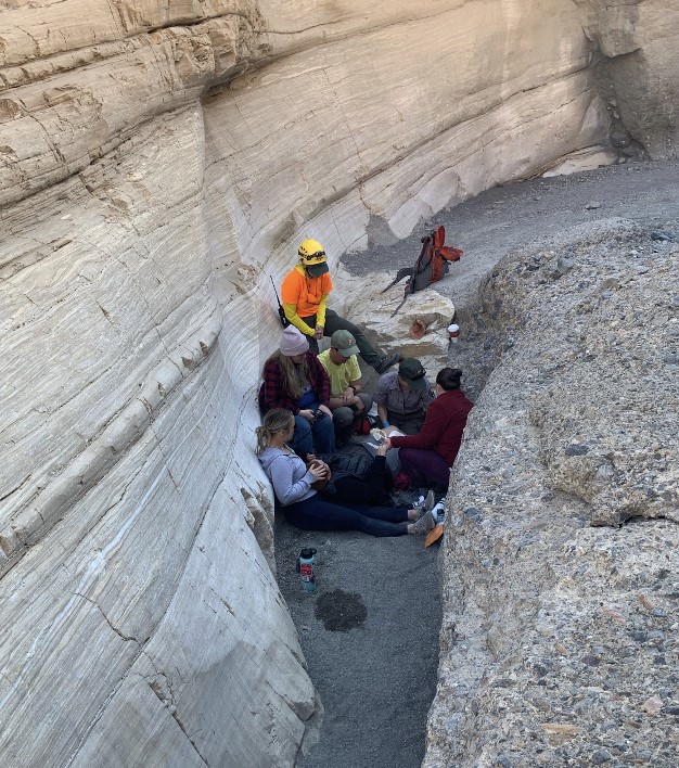 Six people sit around a woman lying on her back in a narrow canyon lined with polished tan wall on the left and a gray conglomerate on the right.