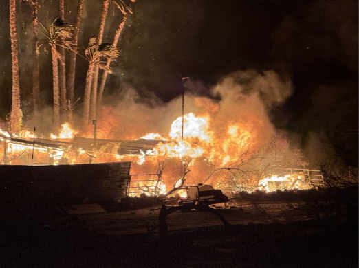 A structure engulfed in intense flames at night, with a large fireball and thick smoke billowing upward. Palm trees are silhouetted against the fire on the left, their fronds glowing orange from the blaze. Dark foreground debris and a fence are visible in