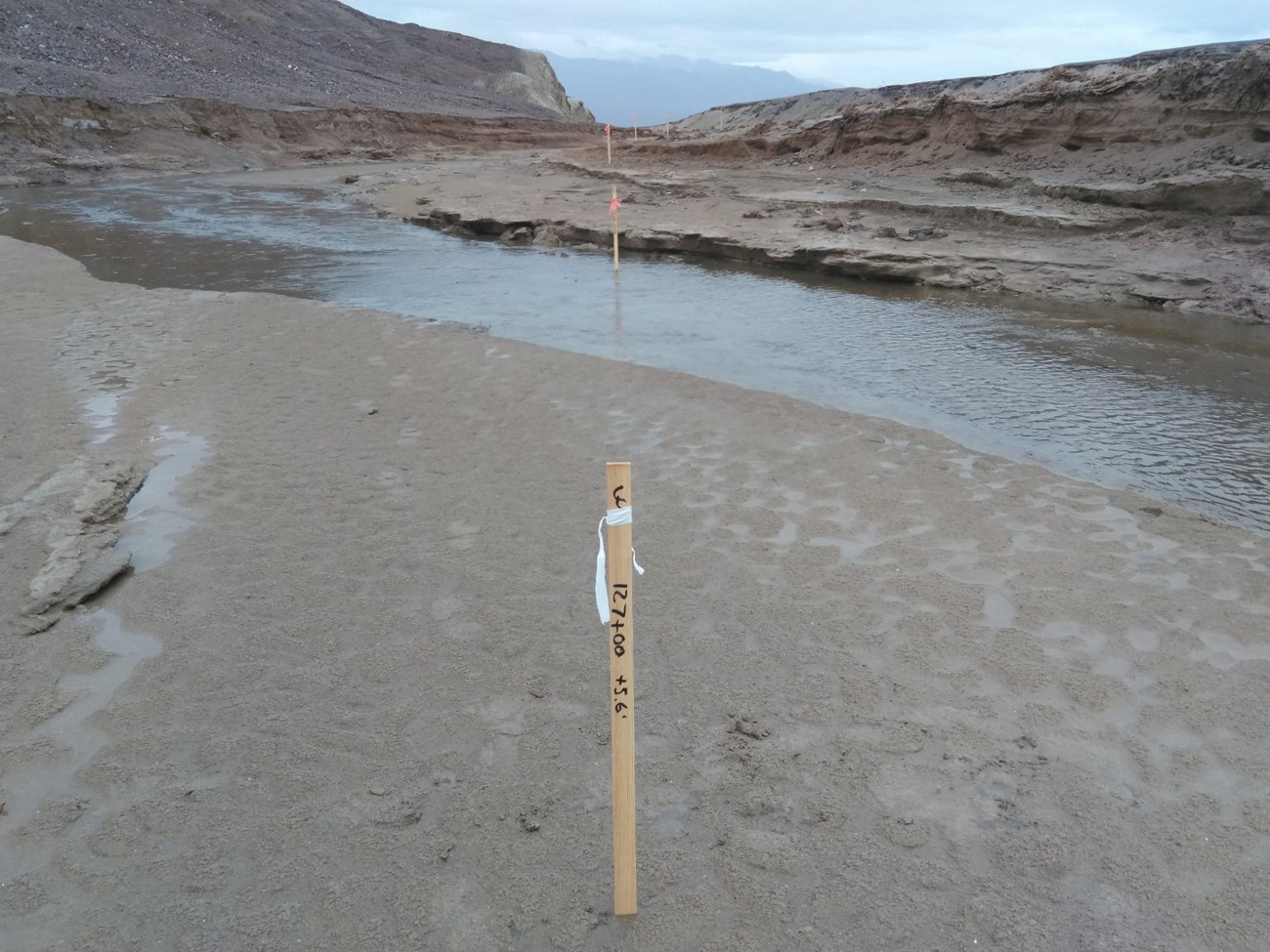 Salt Creek alignment staking 2024-02-05, FHWA A wooden stake is in the ground among a desert landscape that has a shallow stream running through the low hills visibly cut from erosion.
