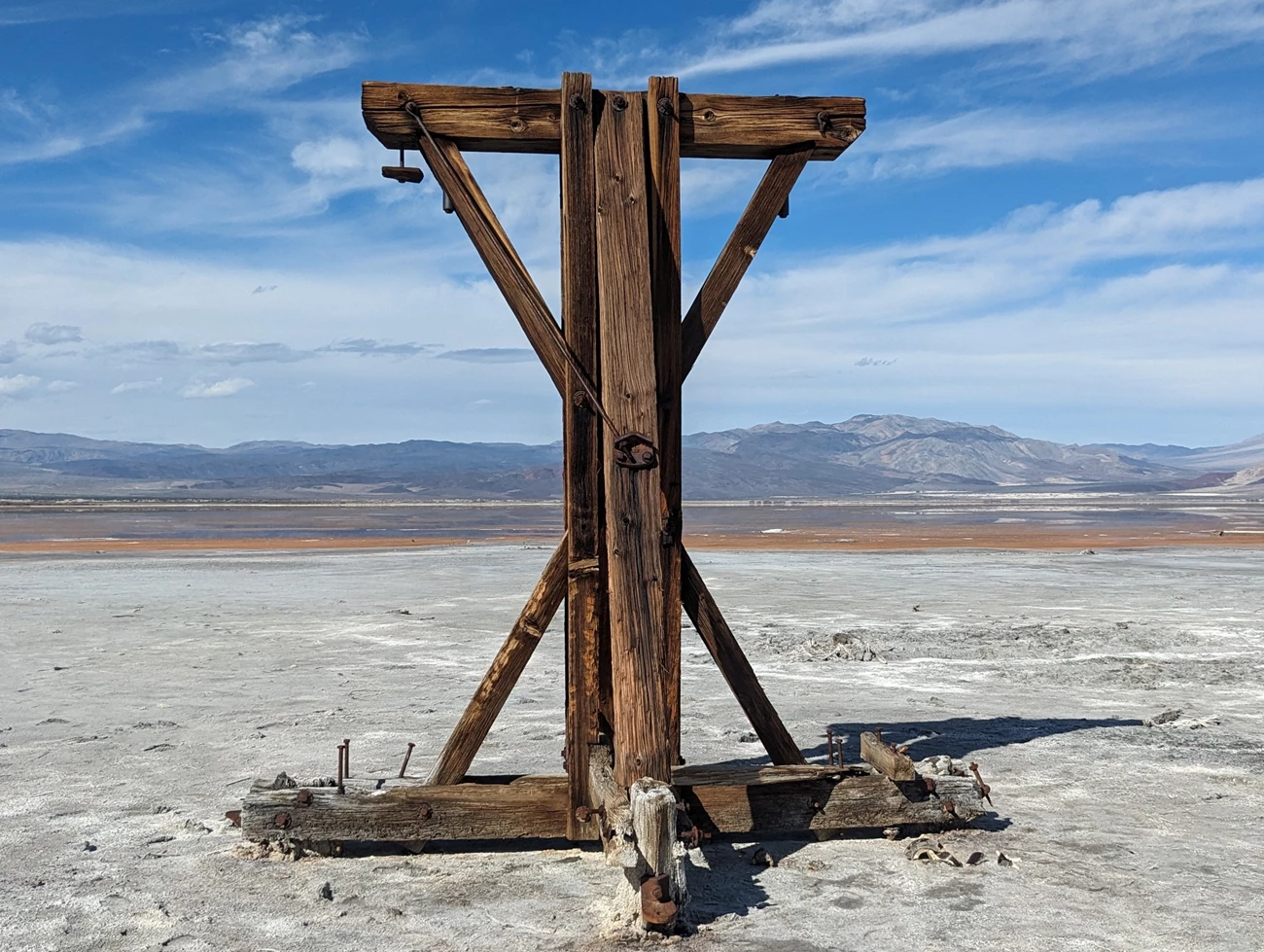 Wooden structure stands upright on flat white ground. A shallow lake and desert mountains are in the background.