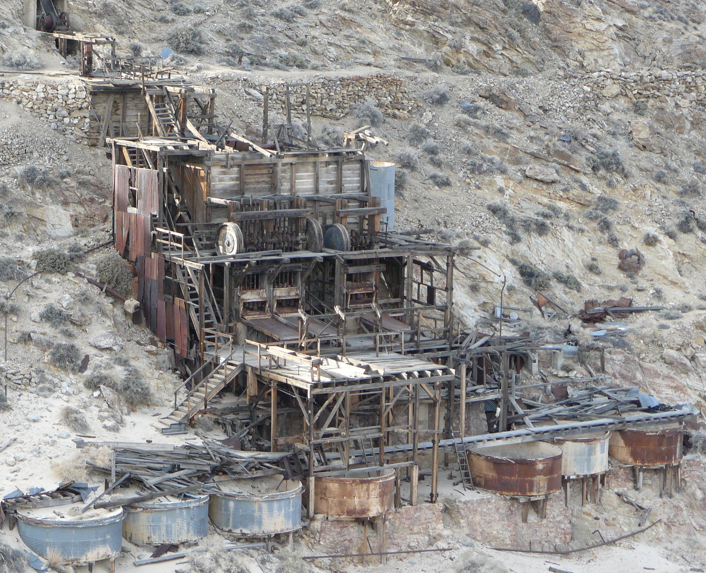 Seven large round metal tanks sit below a multi-story wood and metal structure built on a desert hillside.