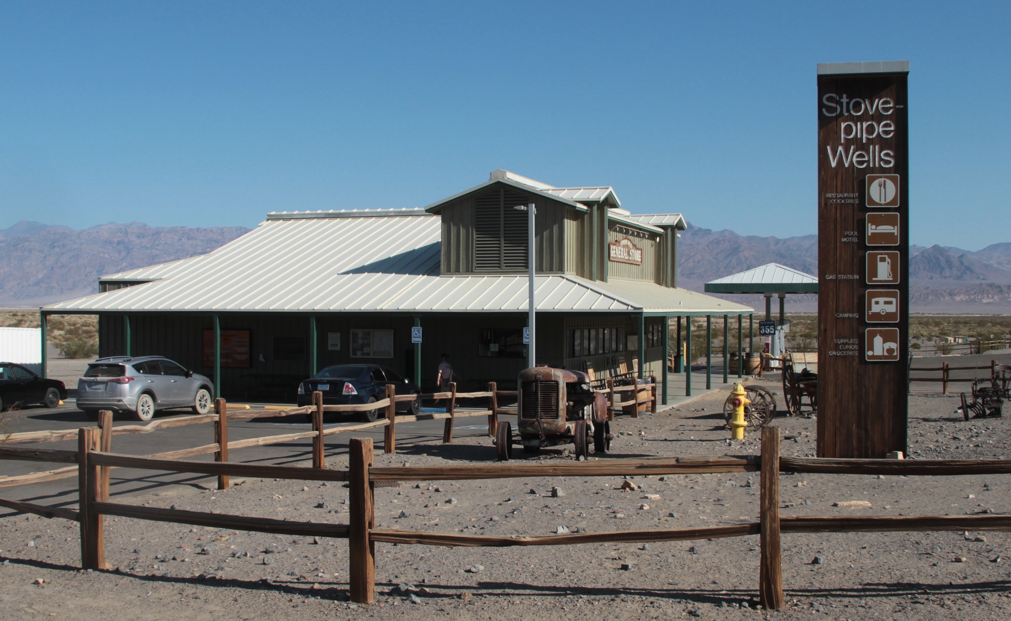 A wooden fence is in the foreground. A store, sign, and cars are in the background.