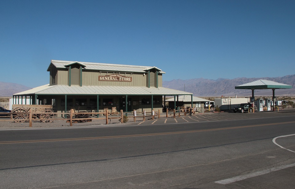 An asphalt road is in the foreground. A sage-color general store is on the left and gas pumps on the right.