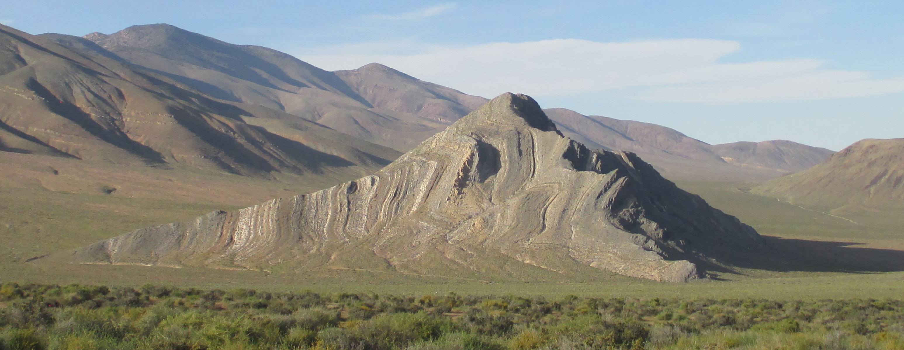 A striped butte stands in the middle of a green valley with shrubs in the foreground and rolling mountains in the background.
