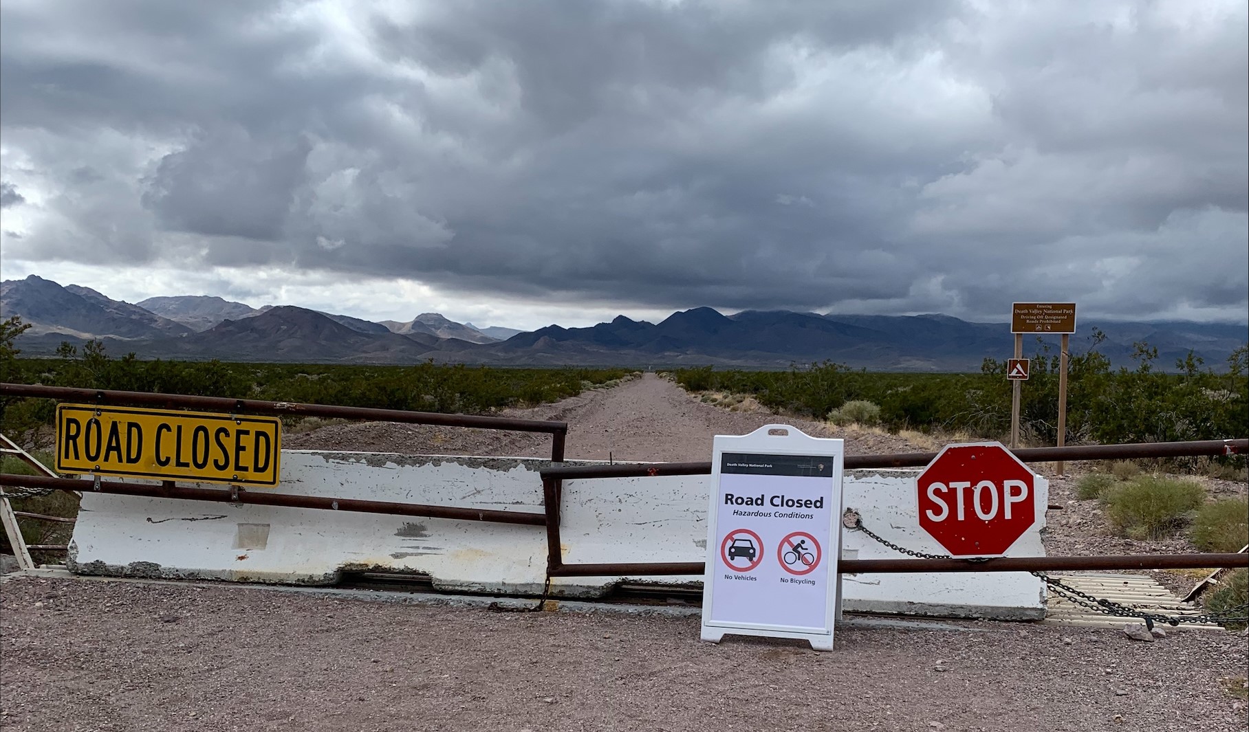 A brown gate, white concrete barriers, yellow "road closed", red "stop" sign, and white "Road Closed" sign with pictograms block a dirt road leading to distant mountains.
