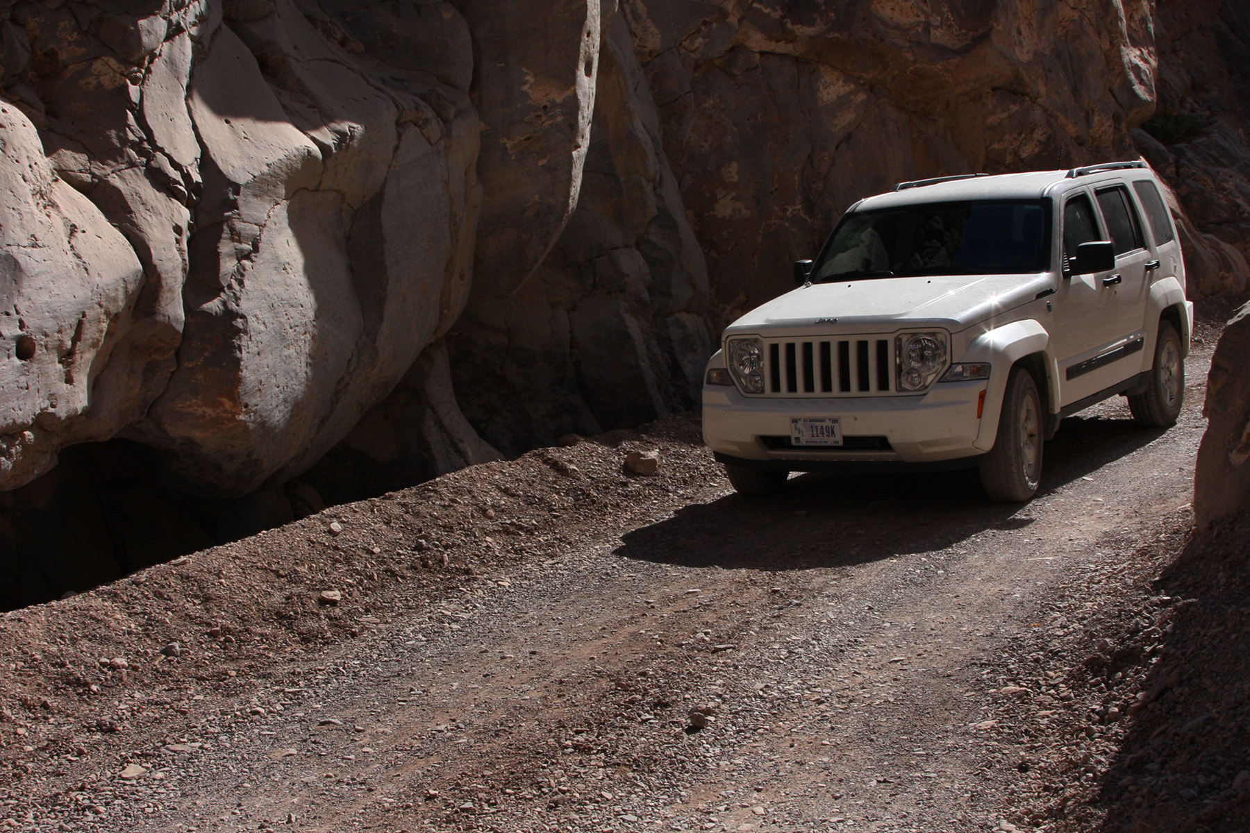 A white Jeep on a one-lane unpaved road between vertical rock walls.