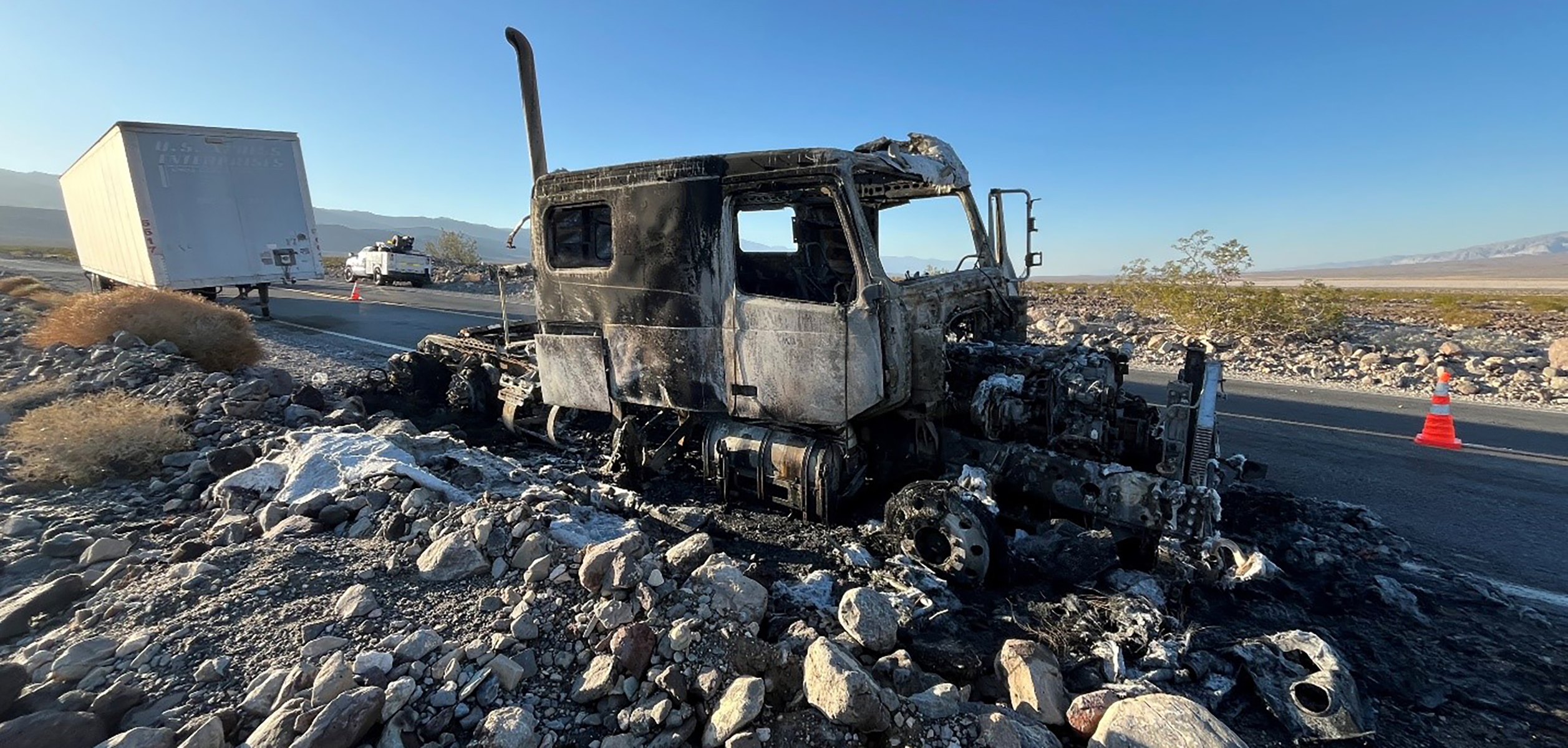 Tractor Trailer Fire_2025 A thoroughly burned truck cab sits abandoned on the rocky shoulder of a rural road under a clear blue sky.