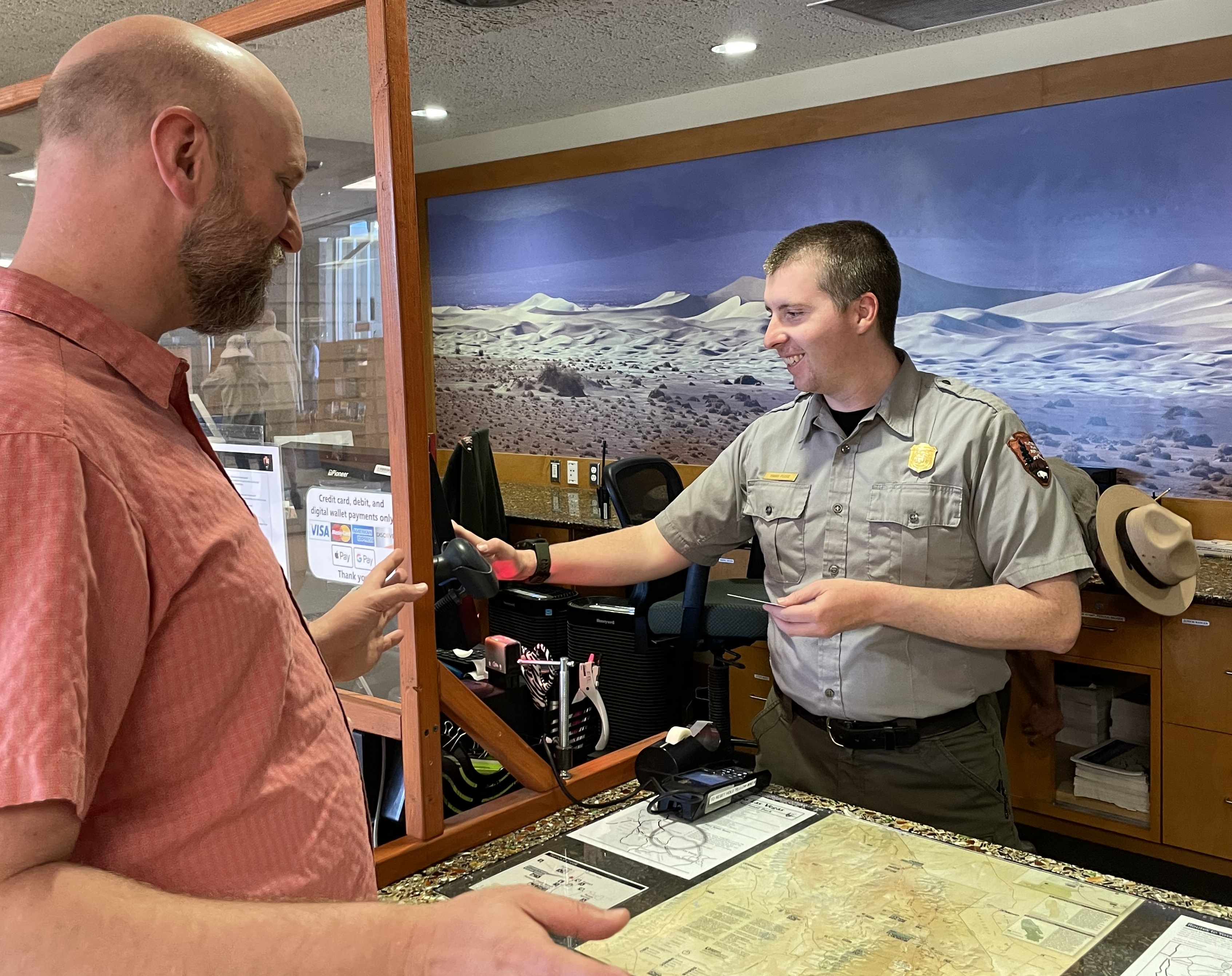 A man in a NPS uniform (gray shirt and green pants) smiles while holding a credit card. Another man in a red shirt stands on the other side of the desk.