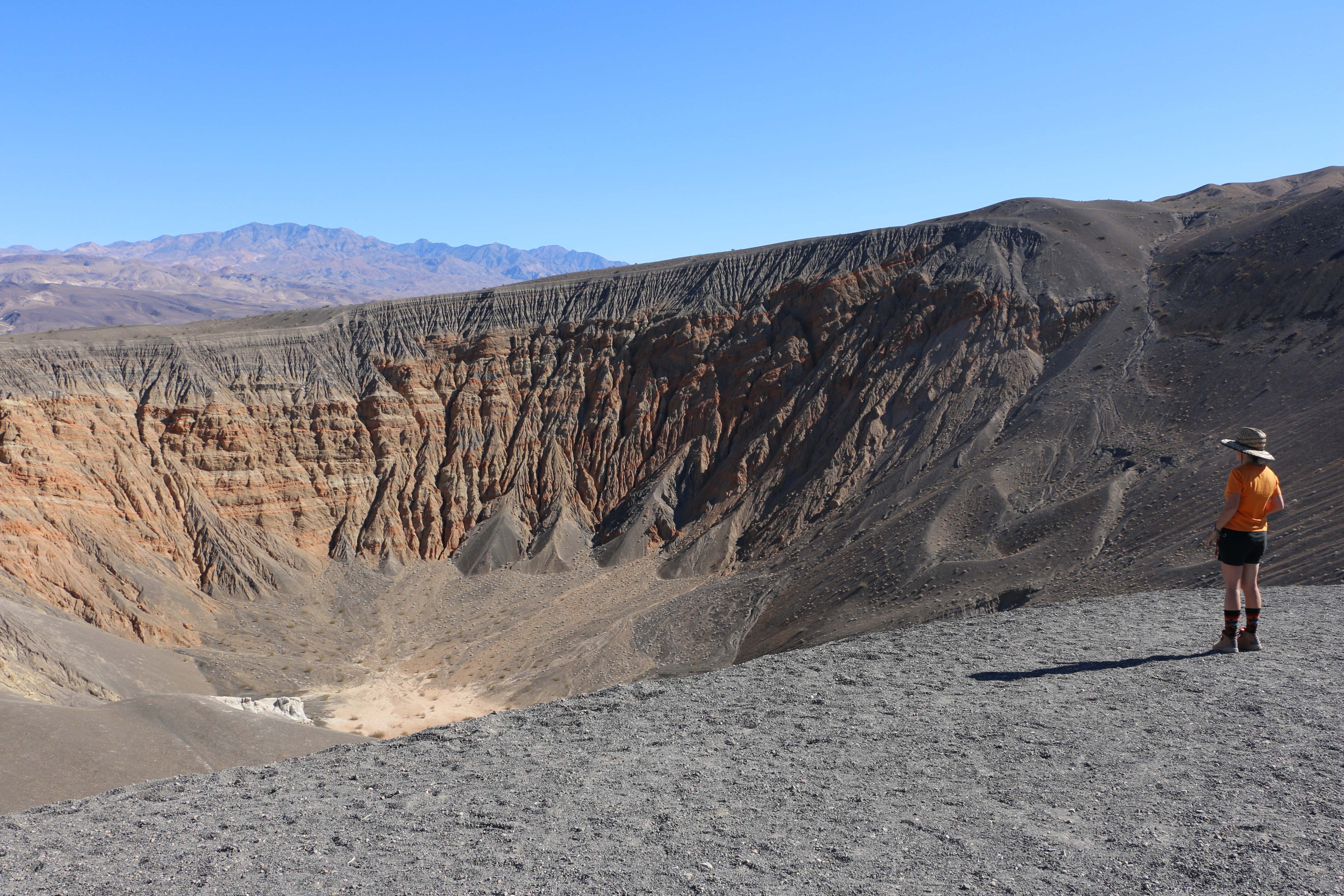 A person wearing an orange shirt and black shorts stands on the right of the photo, looking into a massive crater with gray, orange, and white eroded steep walls.
