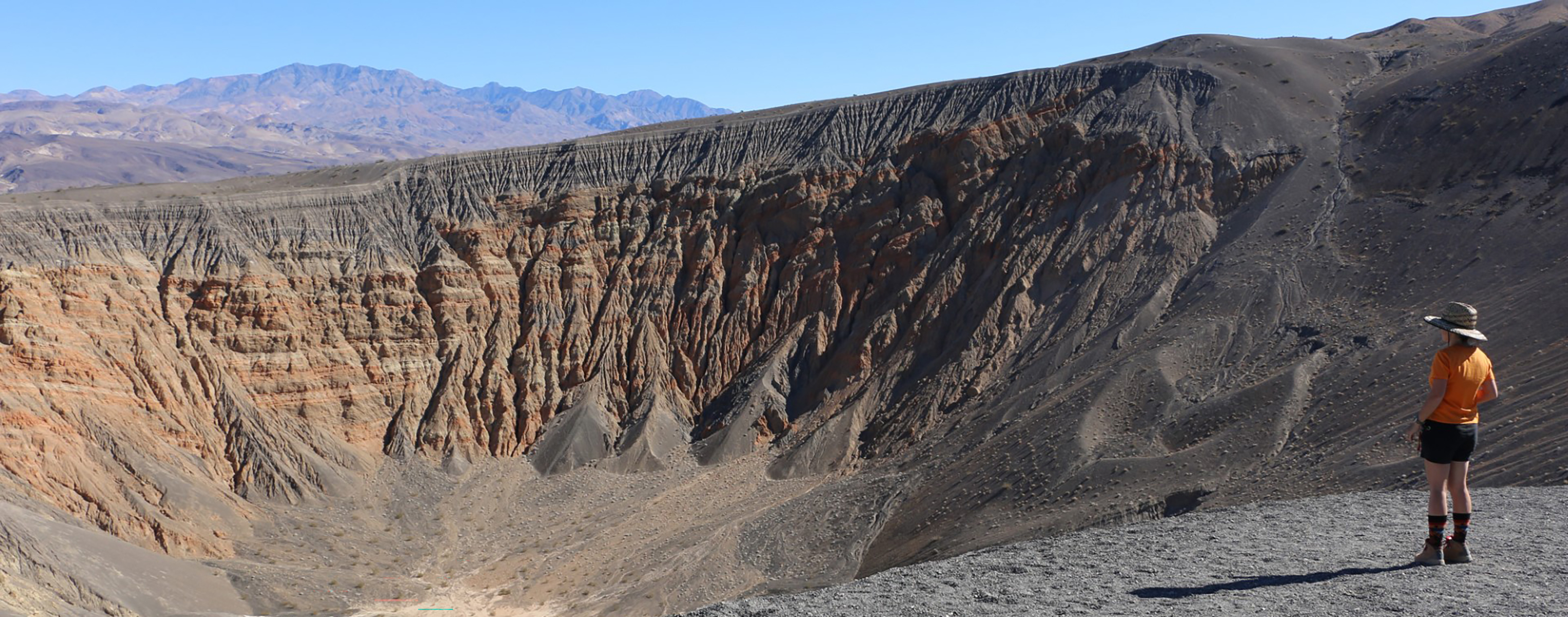 A hiker standing overlooking a large brown, black and orange crate with mountains in the background.