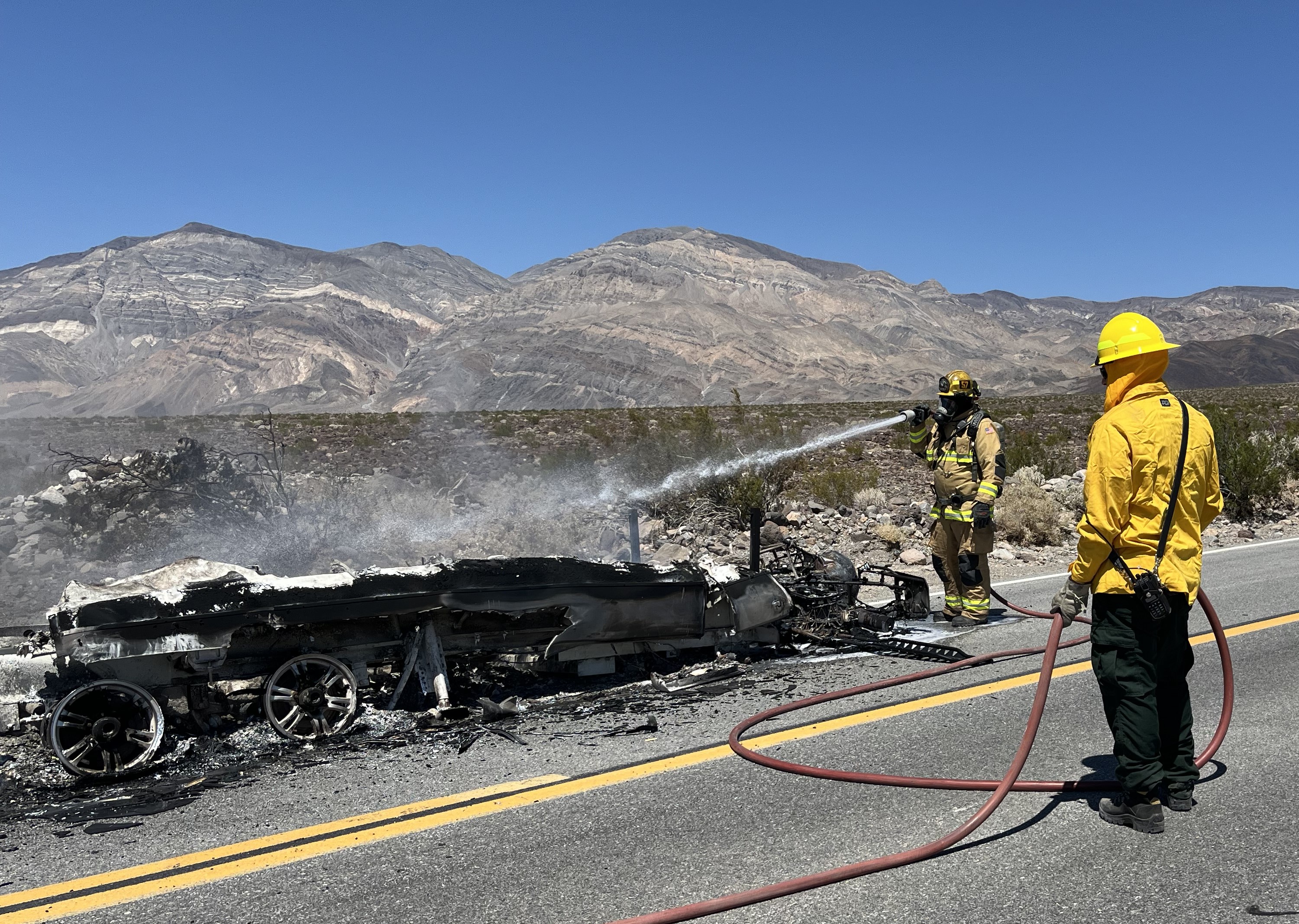 Two people wearing yellow firefighting gear spray water at a burned remnant of a trailer.