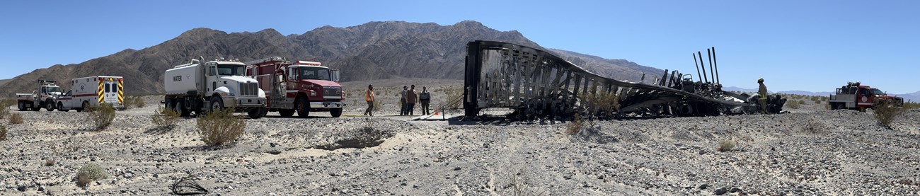 A panoramic image of emergency response vehicles on either side of a black, burned hull of a truck. A barren mountain is in the background.