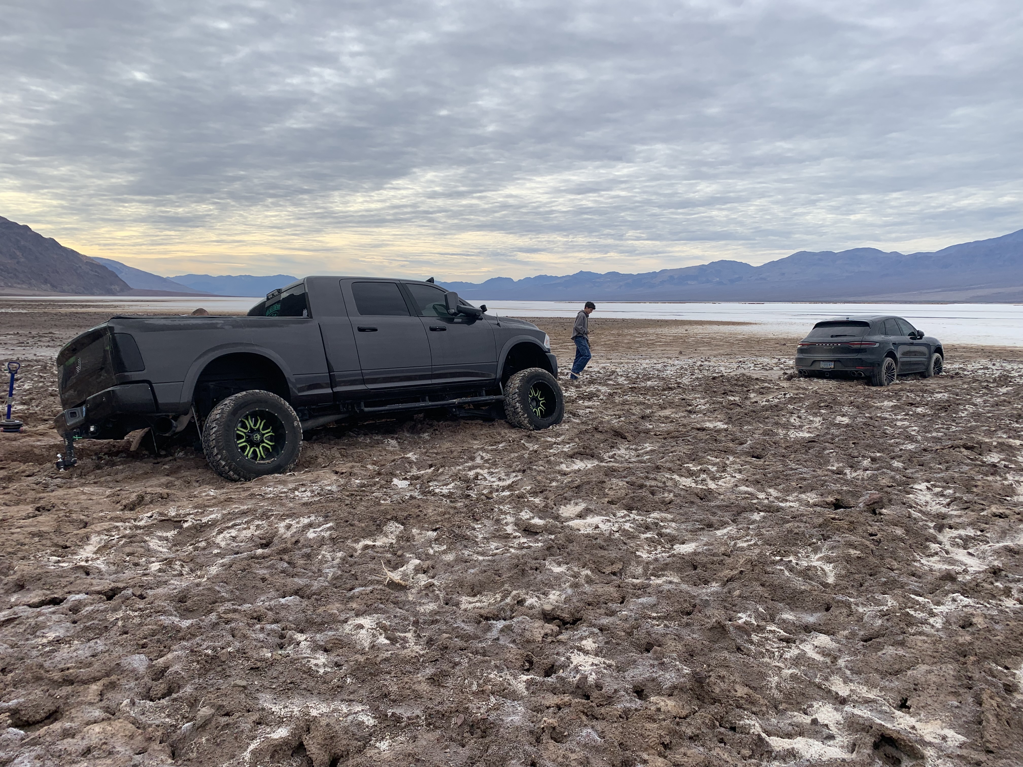 A person walks between a dark gray pickup truck and a black car in an open flat bare landscape
