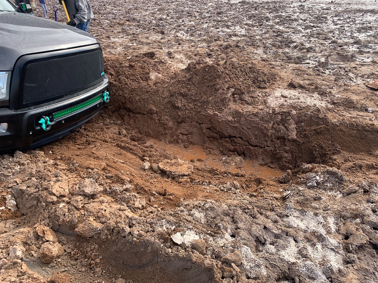 Only the hood and bumper of a gray truck are seen, with a trench in mud in front of it.