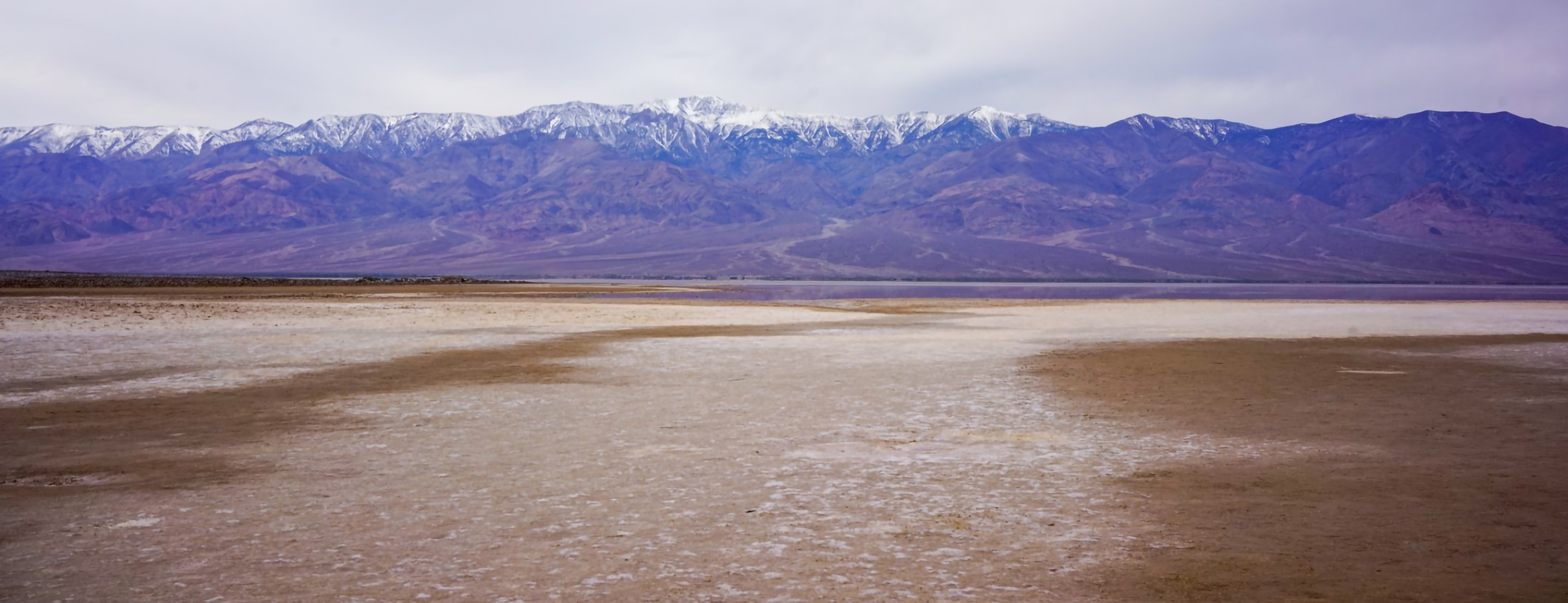 The image shows a flat, dry expanse leading to a sweeping range of purple-blue mountains capped with snow. The open sky above is overcast and muted, creating a cool and subdued atmosphere.