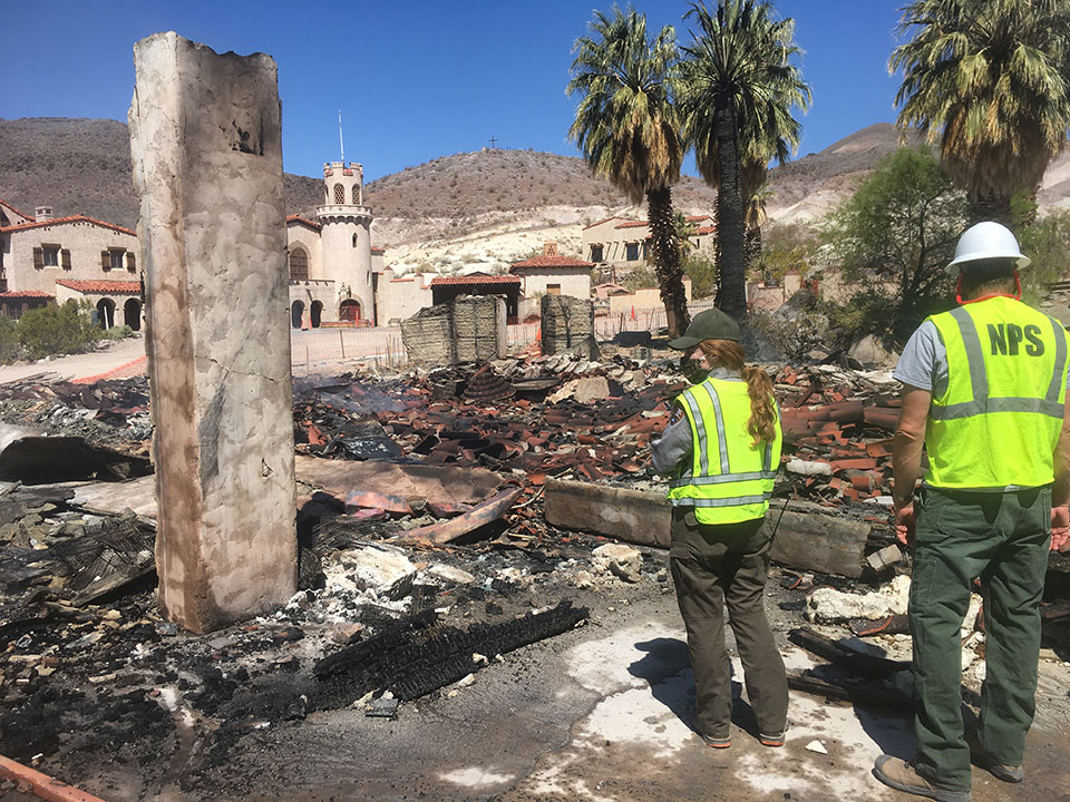 Two rangers wearing high visibility vests and hard hats stand near charred wood and roof tiles with a Spanish-style mansion in the background.