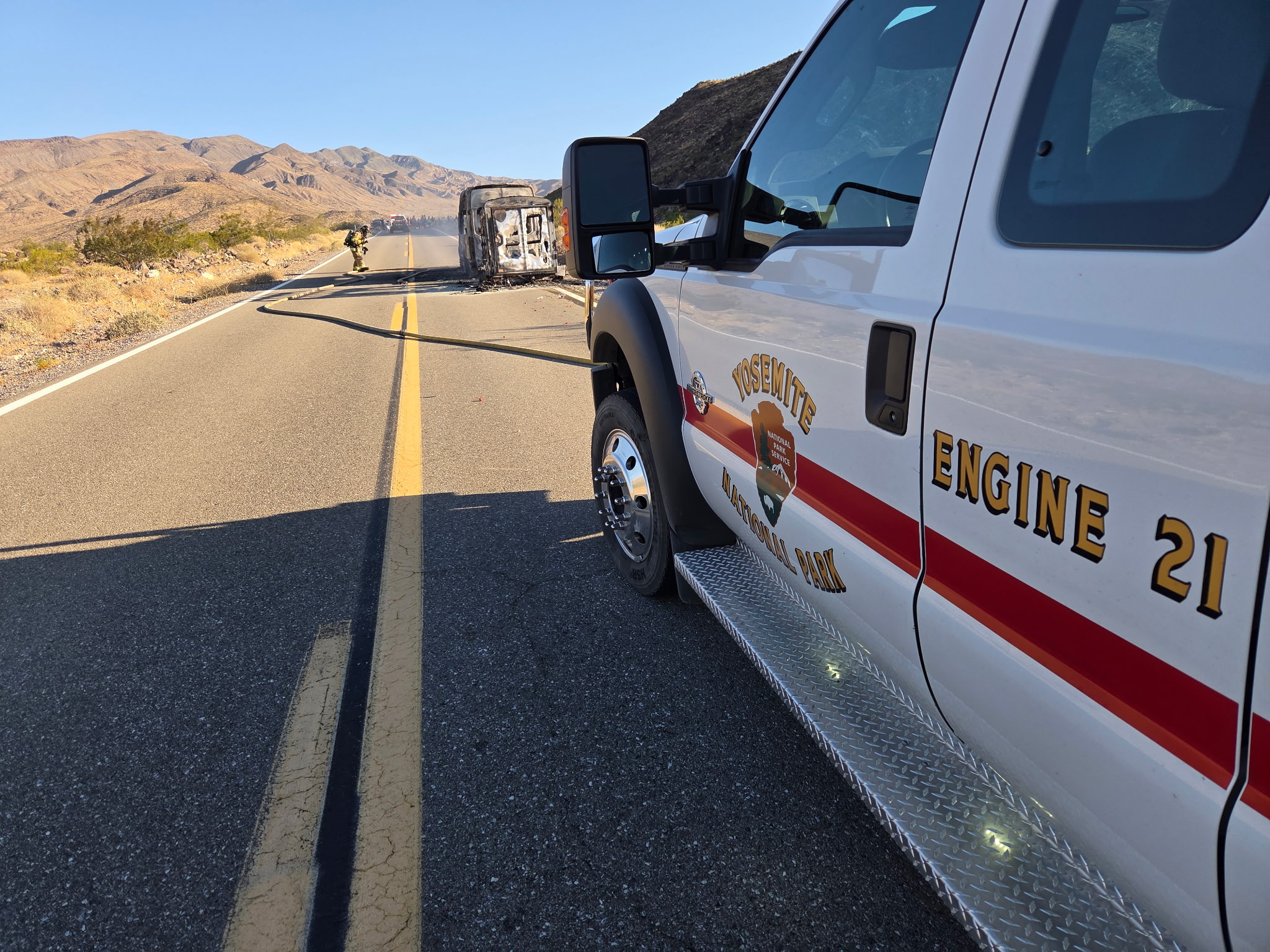 A white vehcile with red stripe is in the foreground. In the background a person in tan clothing holds a firehose near a smoking vehicle.